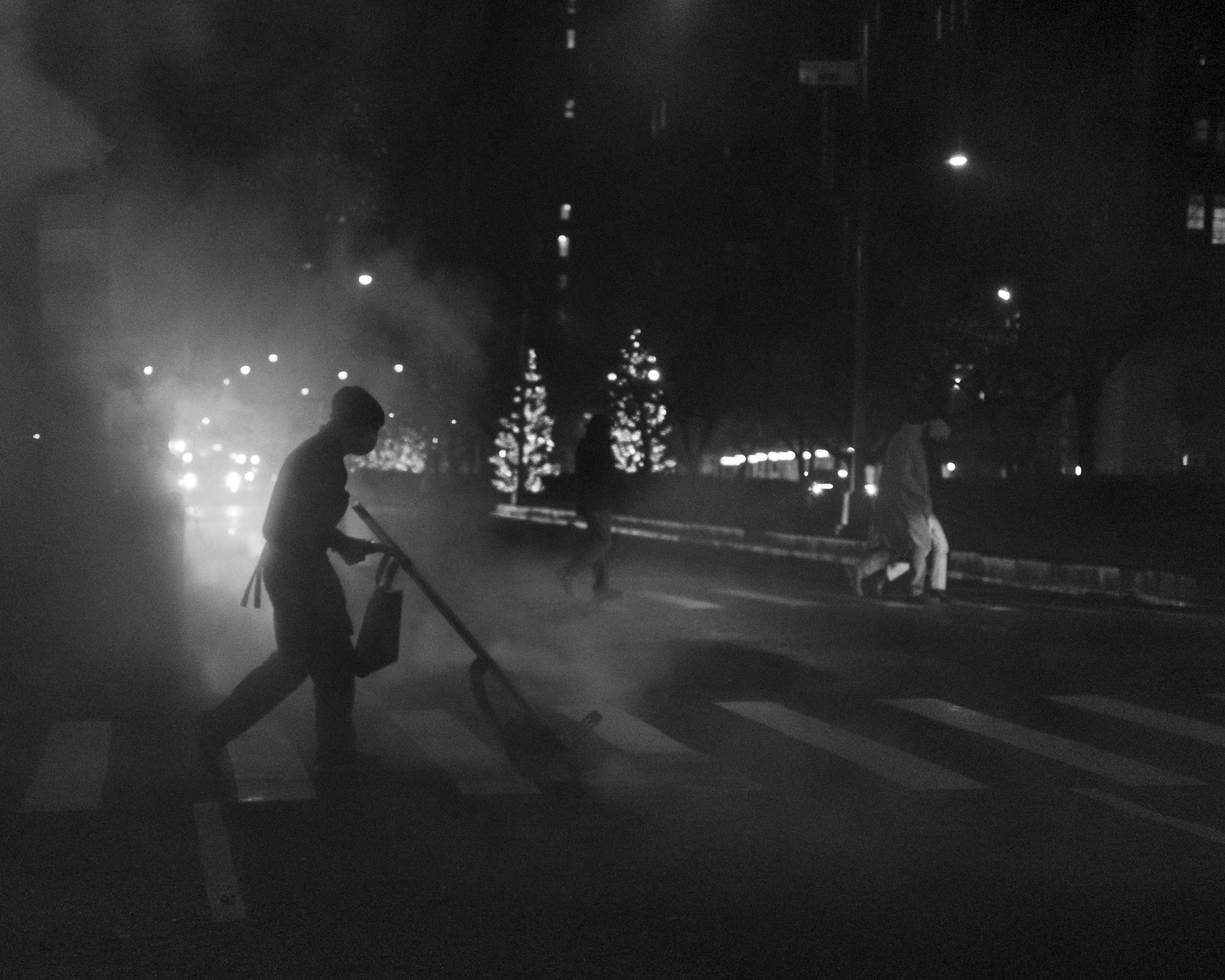 A black and white night photograph of a worker in a knit cap pushing a wide broom across a crosswalk, silhouetted against thick steam lit by headlights. Behind them, two pedestrians cross a divided roadway past trees strung with lights.