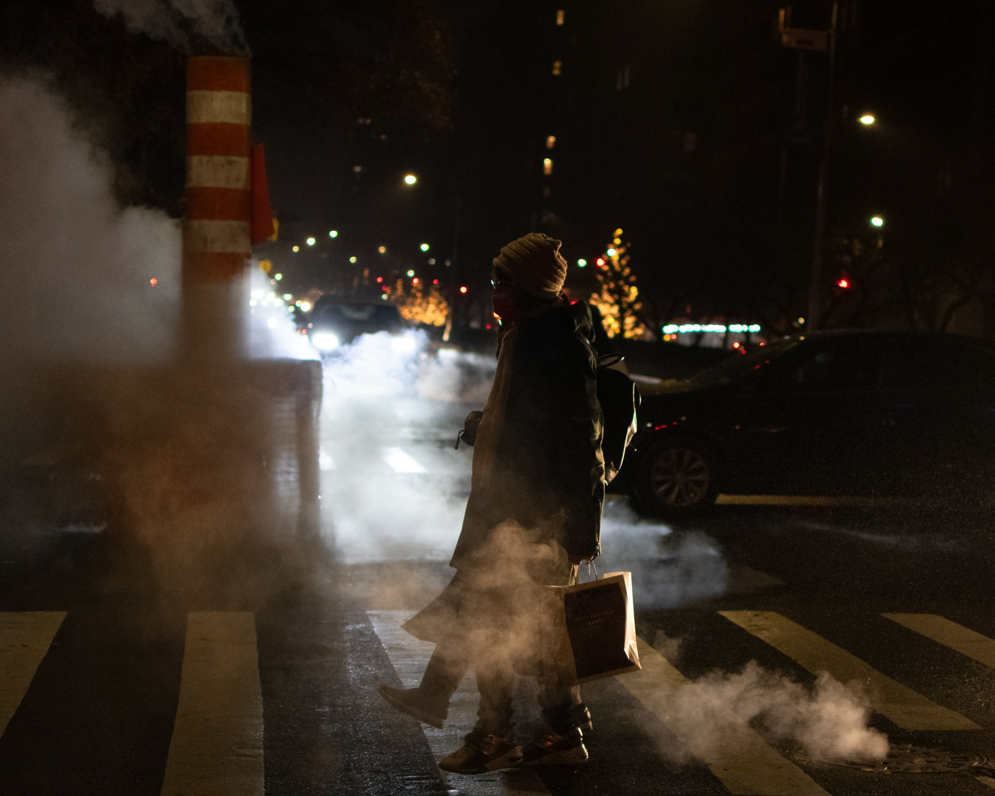 At night, a person in a knit hat, face mask, long dark coat and backpack crosses a wet city street, carrying a paper shopping bag while steam pours from a striped orange and white vent stack and a manhole beside the crosswalk.