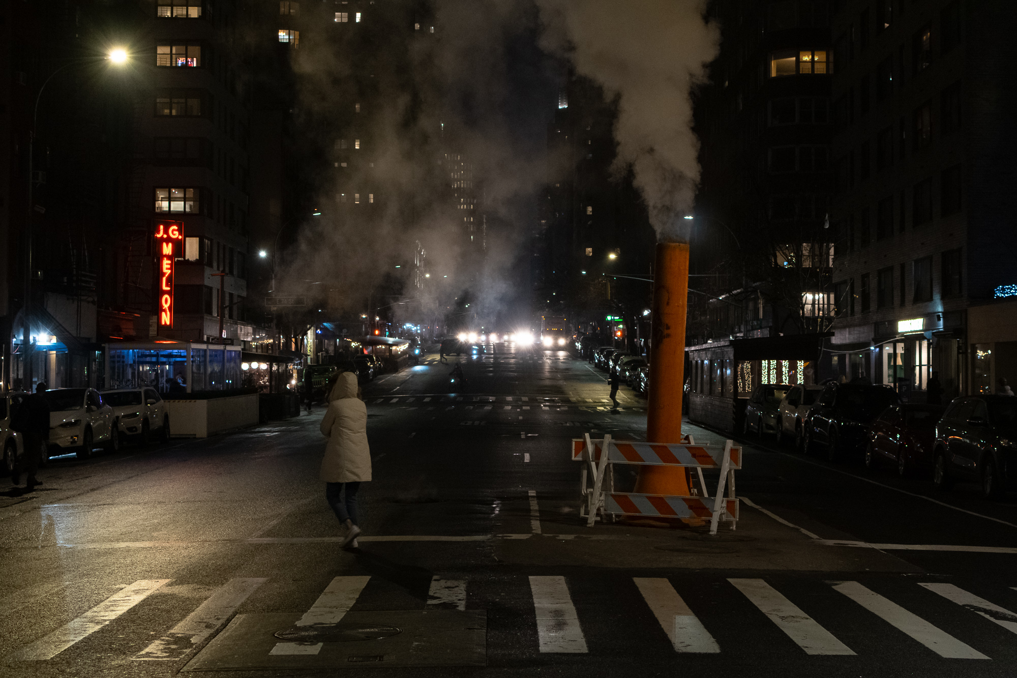 A night street scene in Manhattan where a tall orange steam vent pipe ringed by a striped barricade releases a thick plume into the dark sky, while a person in a pale hooded parka crosses an empty avenue. The red neon J.G. Melon sign glows on a building at left and oncoming headlights shine far down the block.