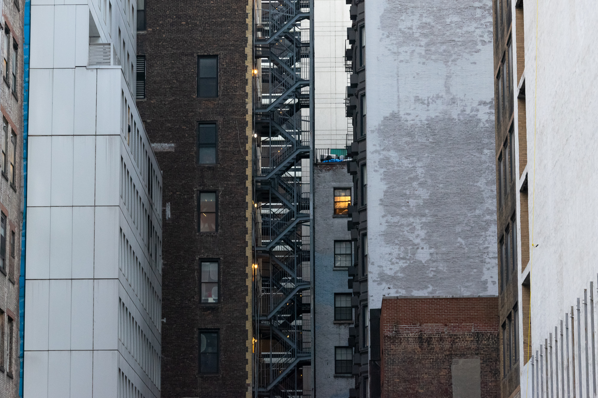 A tightly compressed view down a narrow Manhattan lightwell, where a steel fire escape switchbacks up the full height of a dark brick tenement wall, flanked by a white paneled office facade on the left and a painted-over brick party wall with a single lit window on the right.