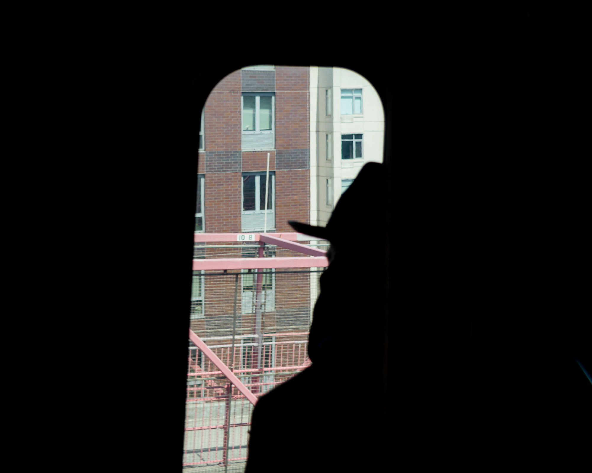 A person's head and shoulders appear in near-total black silhouette against an arch-topped window, through which a brick and cream apartment building is visible above a salmon-pink fire escape stair with mesh railings. A small sign on the railing reads 10 B.