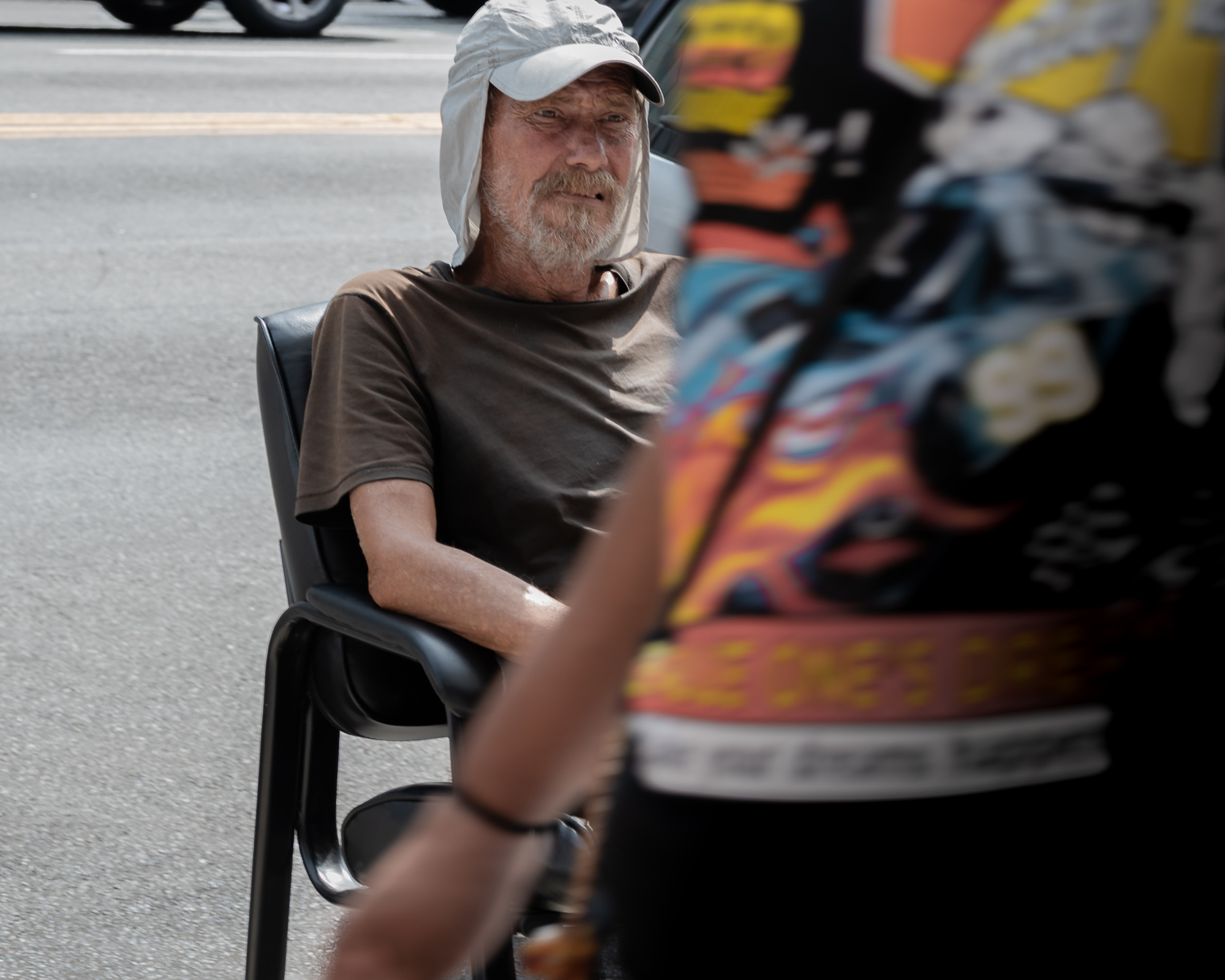 An older bearded man in a khaki cap with a fabric neck flap and a brown T-shirt sits in a black plastic chair at the edge of a street, looking toward the camera. To his right a van covered in bright airbrushed graffiti fills the frame, and a blurred arm with a wristwatch passes close to the lens in the foreground.