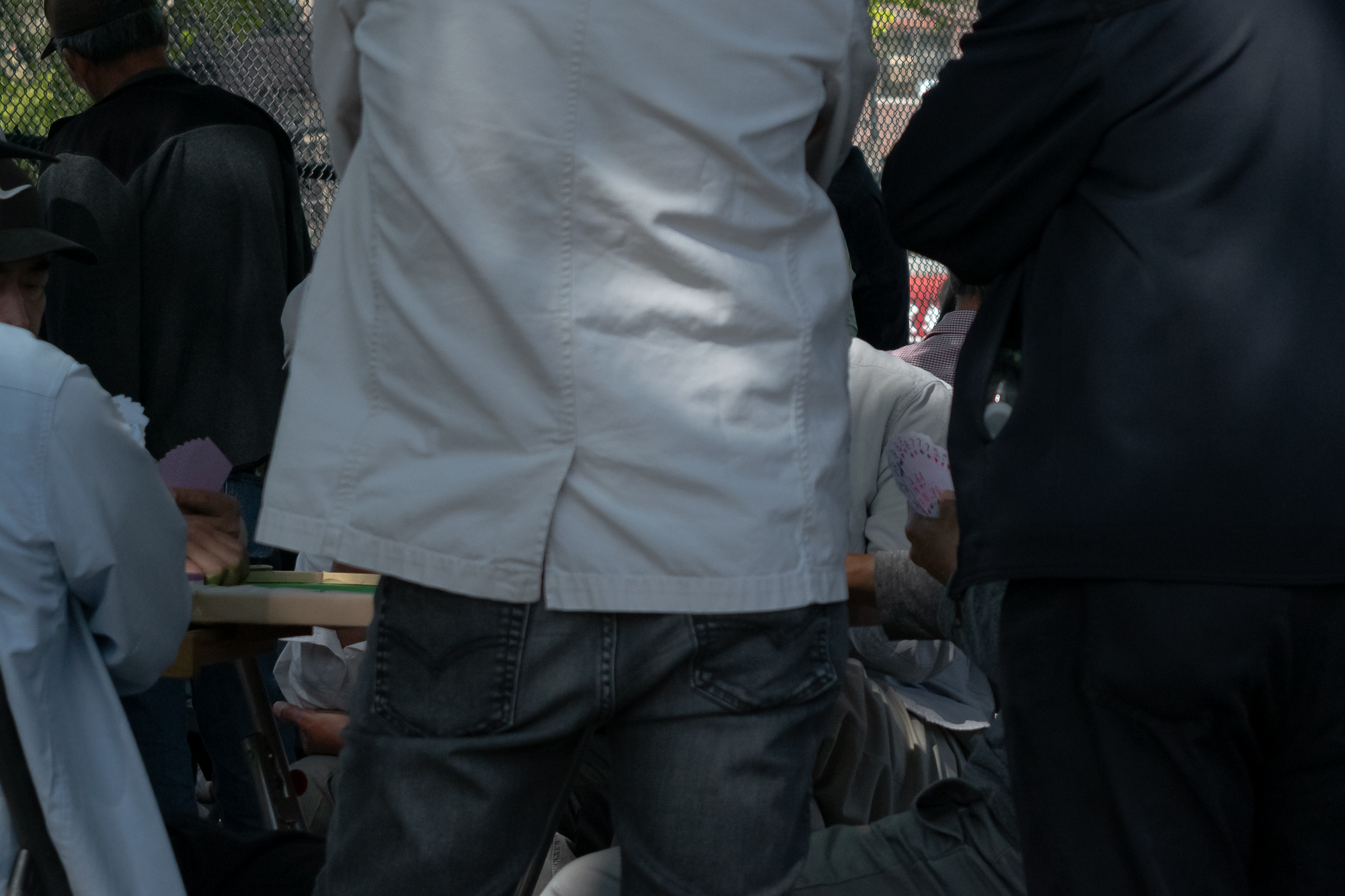 A standing man's back in a pale white jacket and dark jeans fills the center of the frame, blocking most of a folding table where men are seated playing cards. To the right a player holds a fanned hand of playing cards, and a chain-link fence with green foliage rises behind the group.