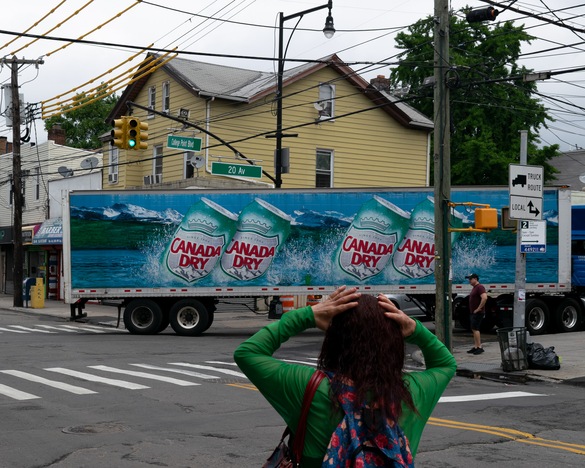 A woman in a green top with both hands on her head stands at a Queens crosswalk, facing a long tractor-trailer painted with giant Canada Dry cans and crashing waves that blocks the intersection of College Point Boulevard and 20 Avenue. Behind the truck sit a yellow clapboard house, tangled overhead wires, a green traffic light and a Truck Route sign.
