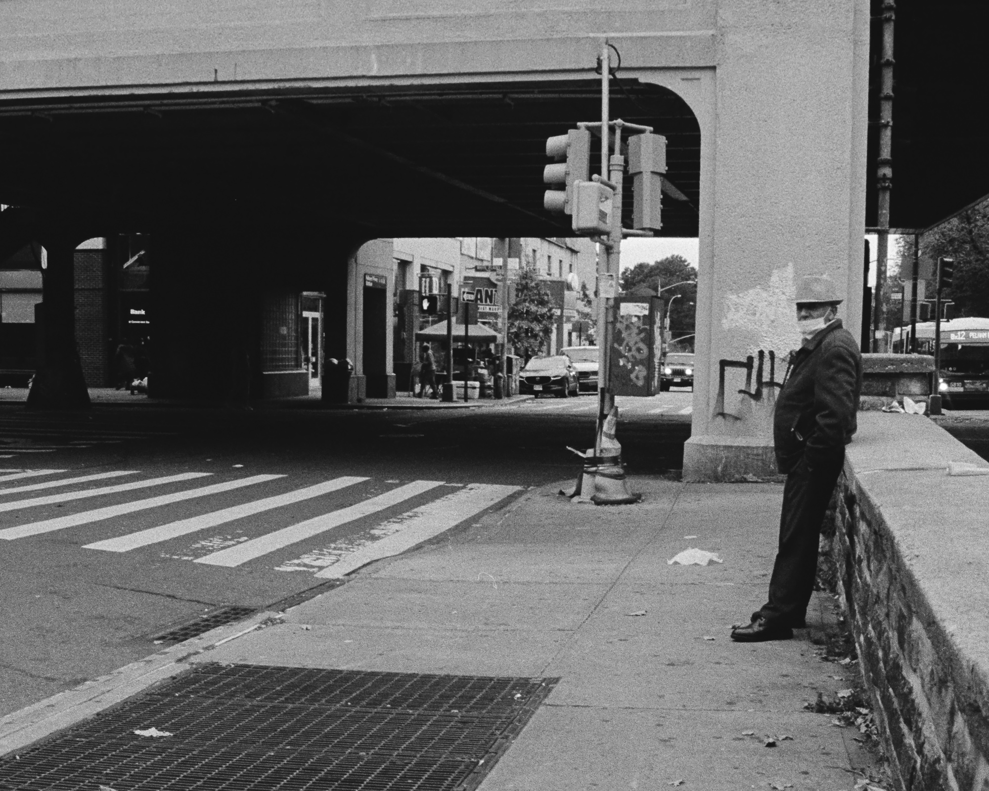 Black and white street photograph of an older man in a fedora, surgical mask and dark jacket leaning against a low stone wall on a sidewalk beneath an elevated train viaduct, turning to look toward the camera. Across the empty crosswalk, a traffic signal, a graffiti-covered utility box, storefronts and a bus sit in the shadow of the overpass.