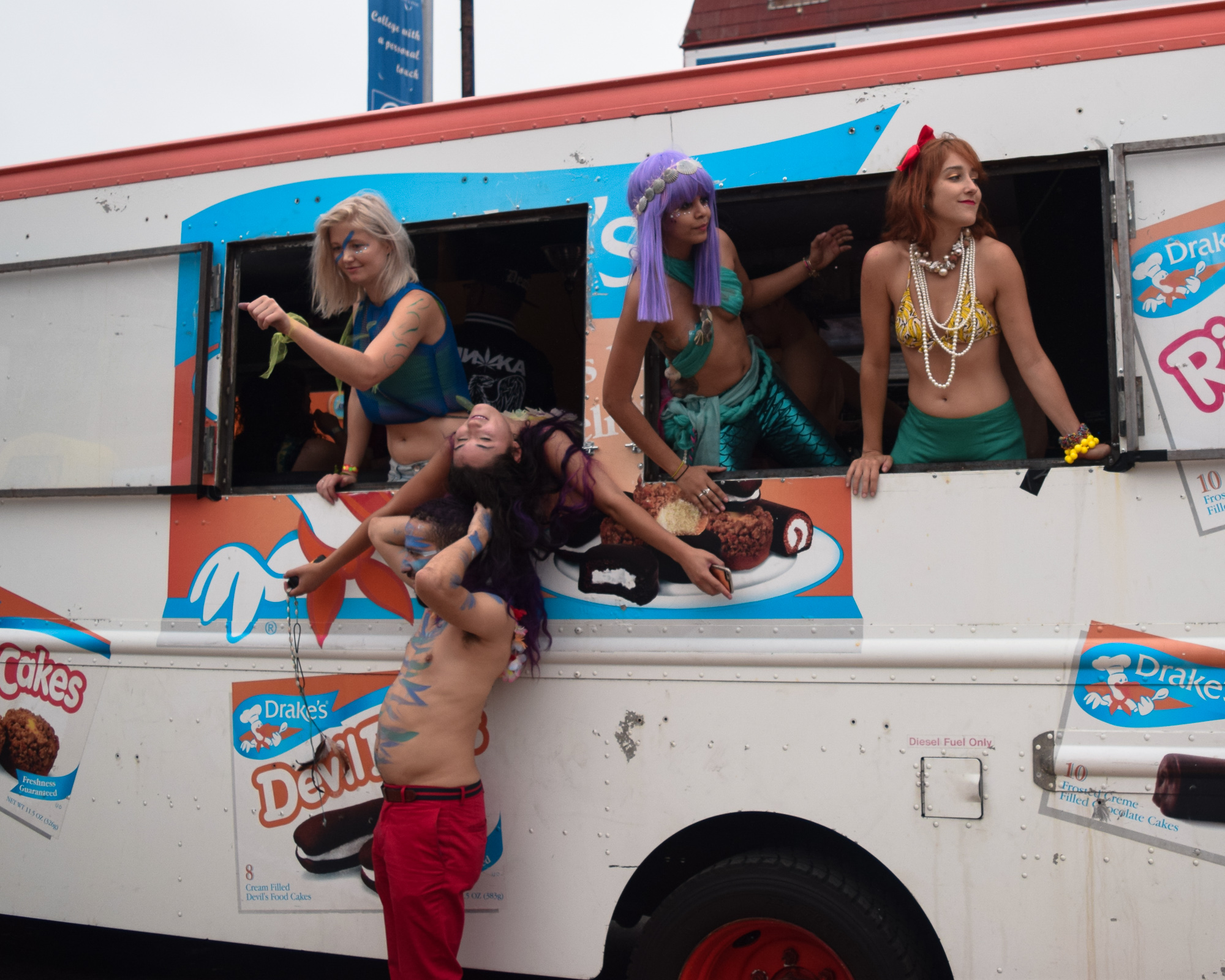 Four costumed performers pose in and around a white delivery truck painted with Drake's Devil Dogs snack cake advertising. Three women in wigs, shells and bikini tops lean out of open side windows while a shirtless man with blue body paint and red trousers carries a woman with purple hair draped across his shoulders.