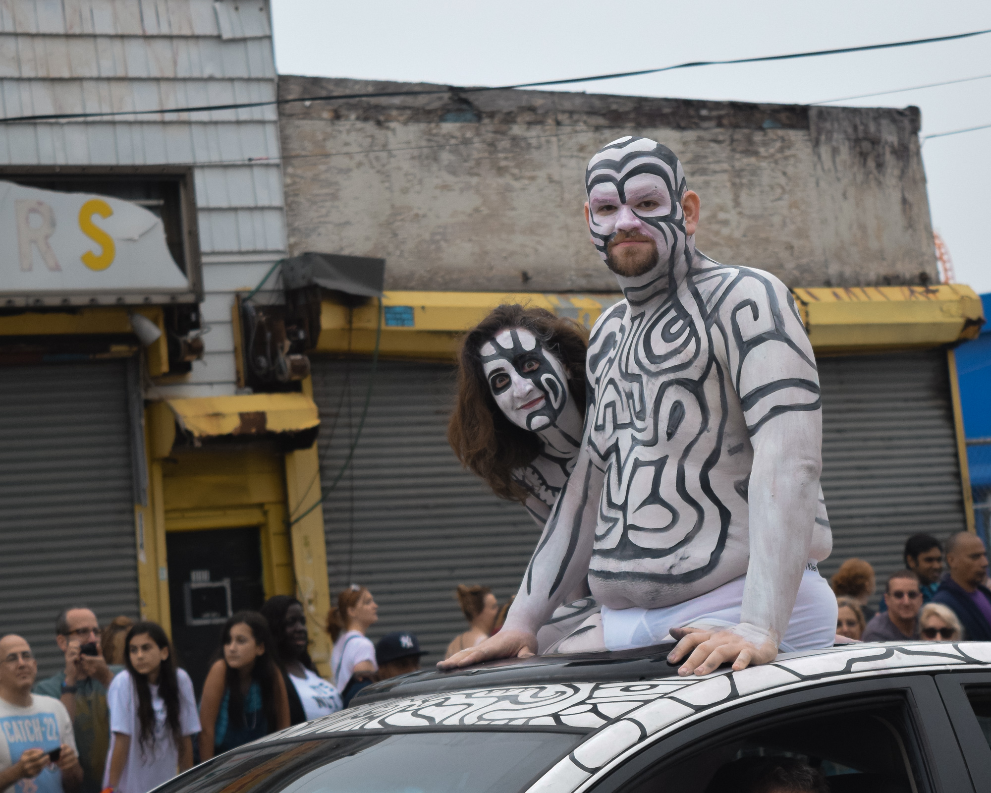 A man and a woman painted head to torso in black and white maze-like patterns kneel on the roof of a car whose paintwork carries the same design, riding past a shuttered storefront with a yellow awning while a crowd watches from the sidewalk.