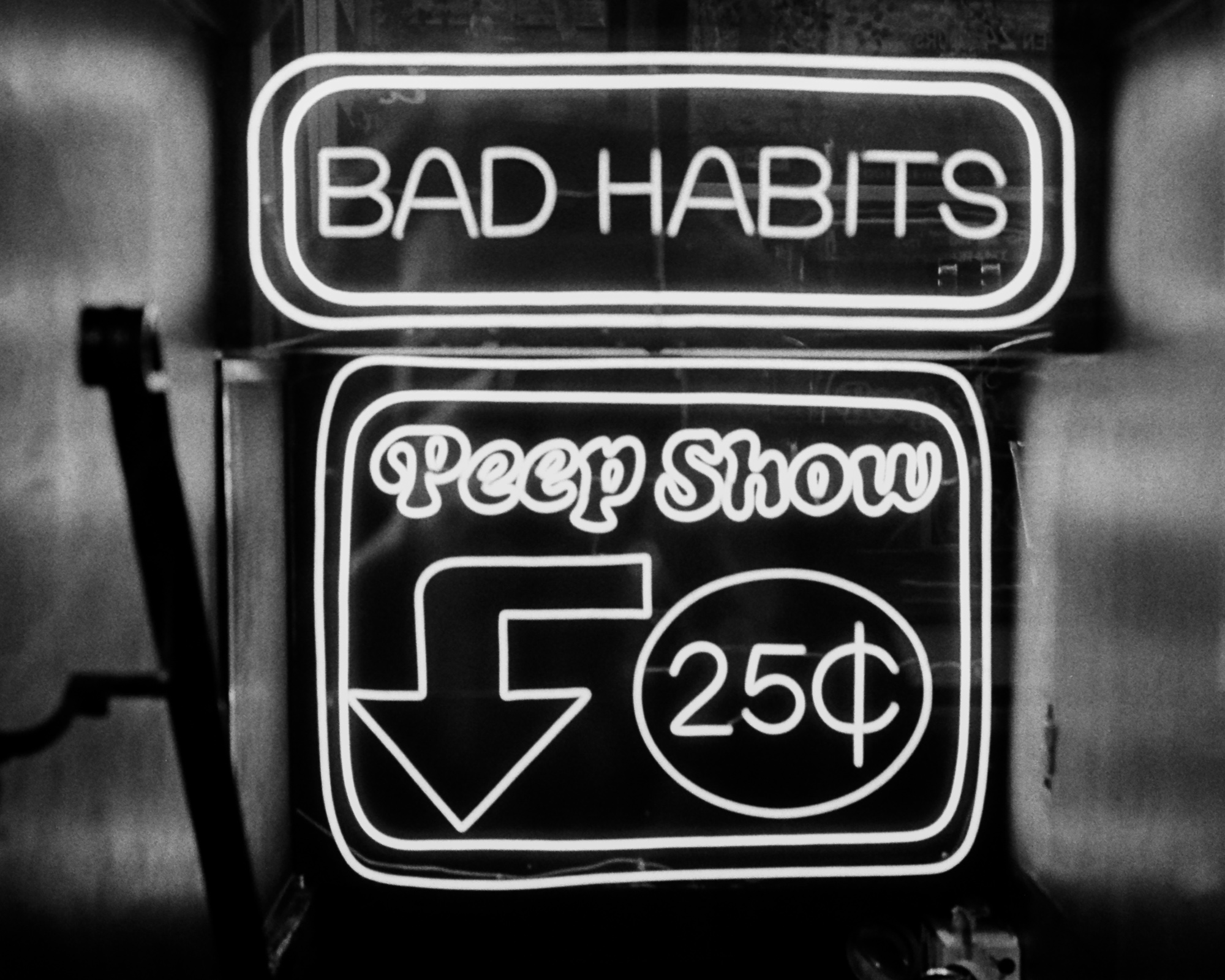 A black and white night photograph of two neon signs in a storefront window. The upper sign reads BAD HABITS in block capitals inside a rounded rectangle; the lower sign reads Peep Show in script above a large downward arrow and a circle enclosing 25 cents.