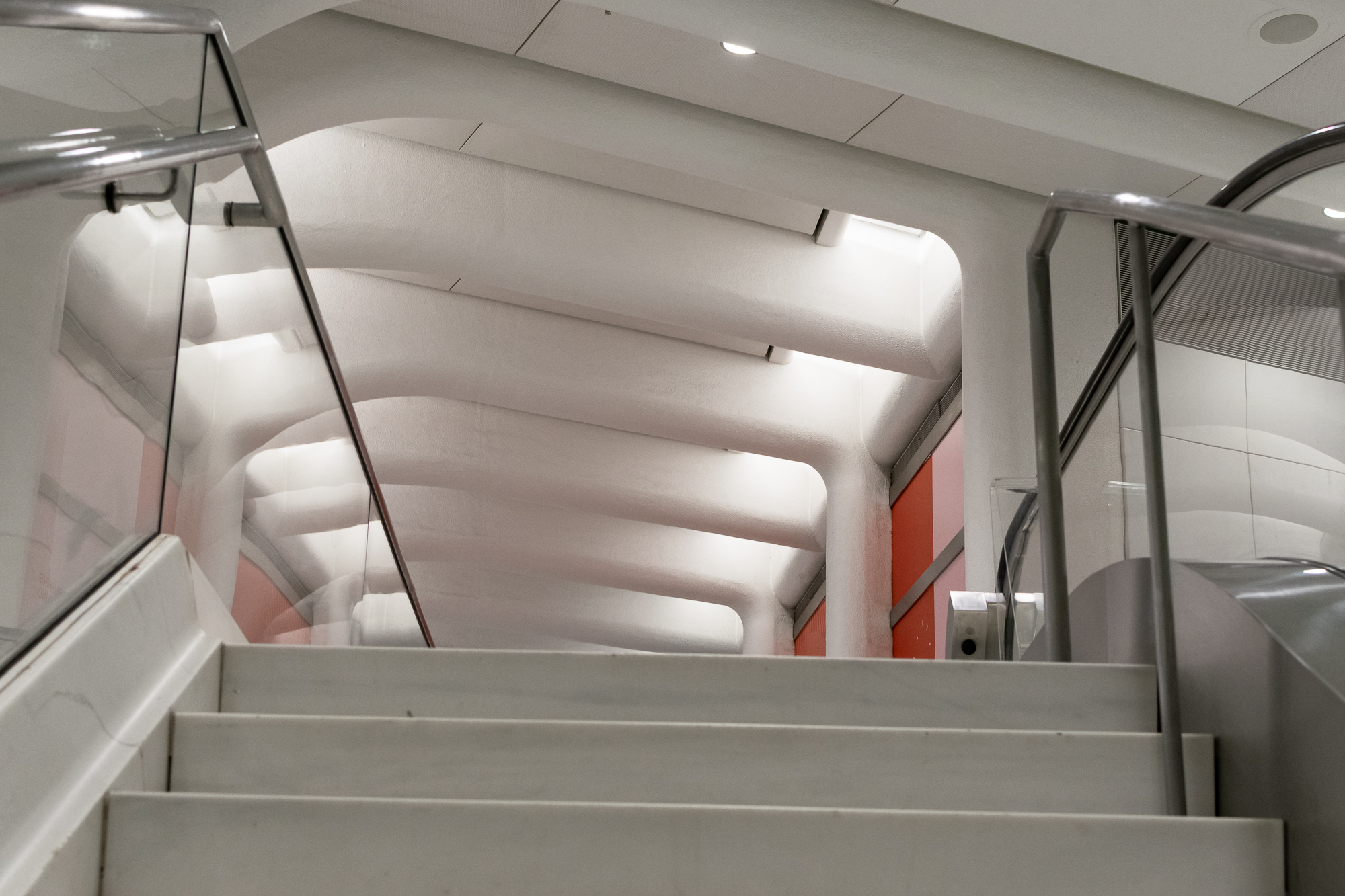 View up a wide concrete stairway toward a white ribbed vaulted ceiling with rounded beams and recessed strip lights, framed by glass panels and tubular steel handrails, with an orange wall panel and escalator edge at right.