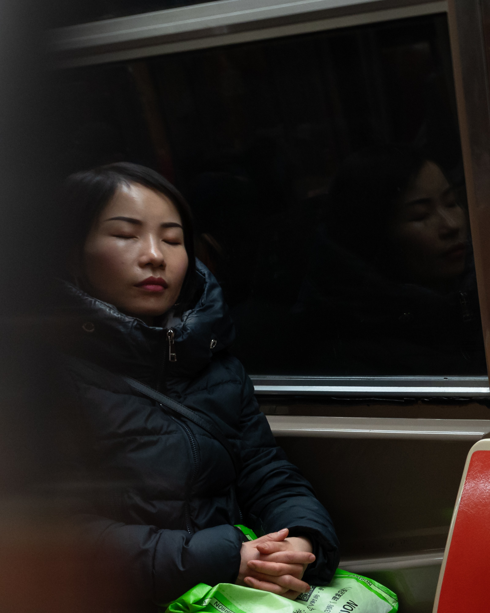 A woman in a black puffer coat sleeps upright on a subway seat, eyes closed and hands folded over a bright green shopping bag in her lap. Her face is reflected in the dark train window beside her, with a red seat edge visible at the right.