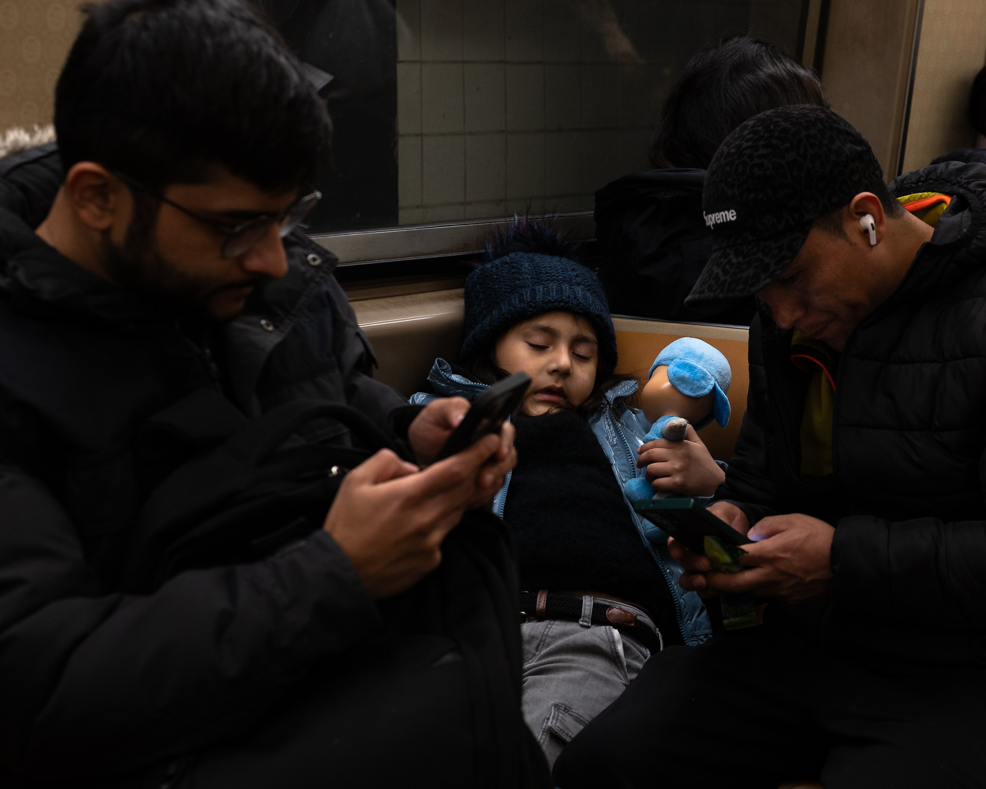 On a subway bench, a child in a navy knit pom hat and pale blue puffer coat sleeps upright holding a blue stuffed animal, while a bearded man in glasses on the left and a man in a Supreme cap with a white earbud on the right both look down at their phones.
