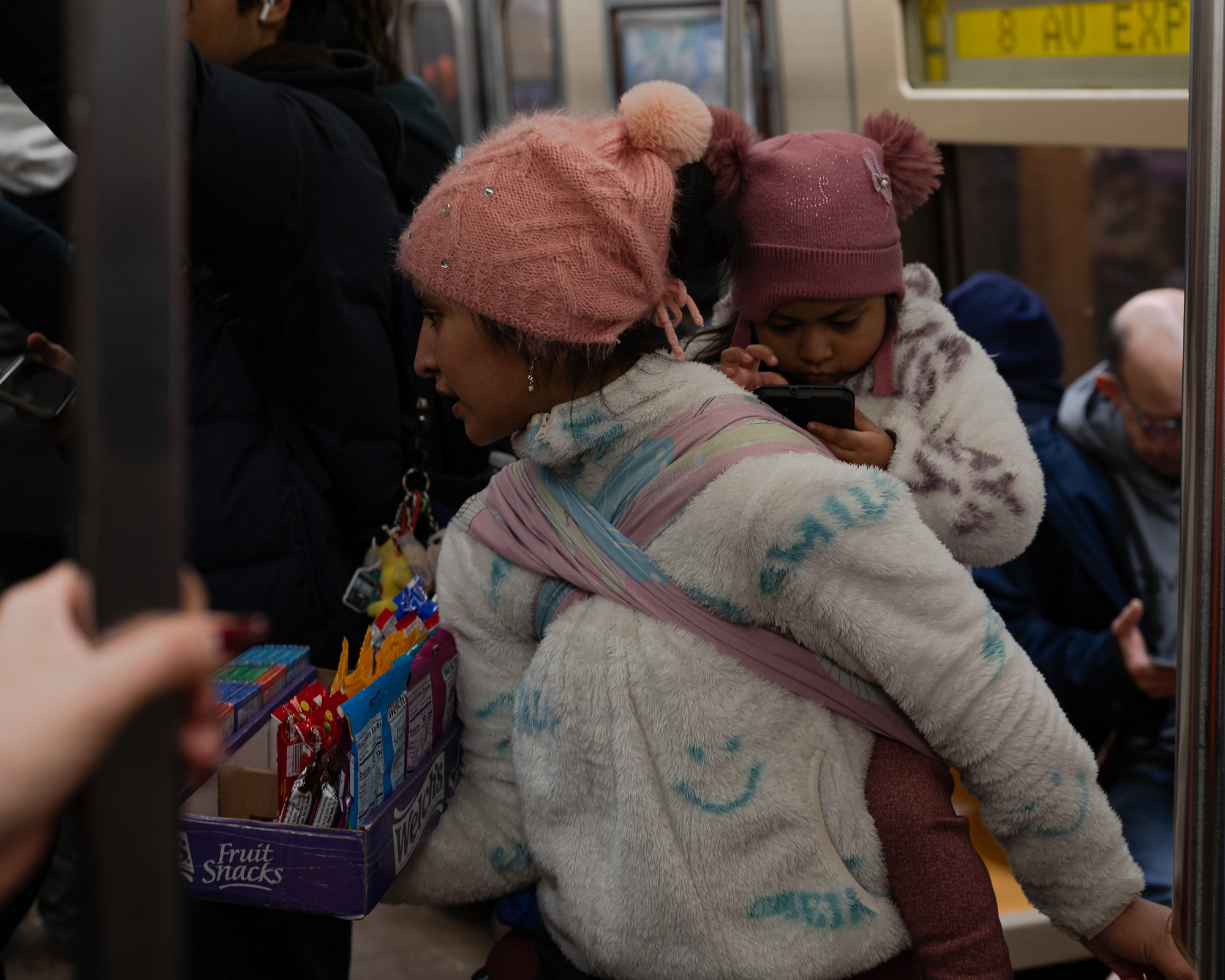 A woman in a pink knit pom-pom hat and cream fleece coat carries a young girl on her back in a wrapped cloth sling, while balancing a Welch's Fruit Snacks box filled with candy and gum against her hip inside a subway car. The child, in a matching rose-colored pom hat, holds a phone close to her face as passengers stand and sit around them.