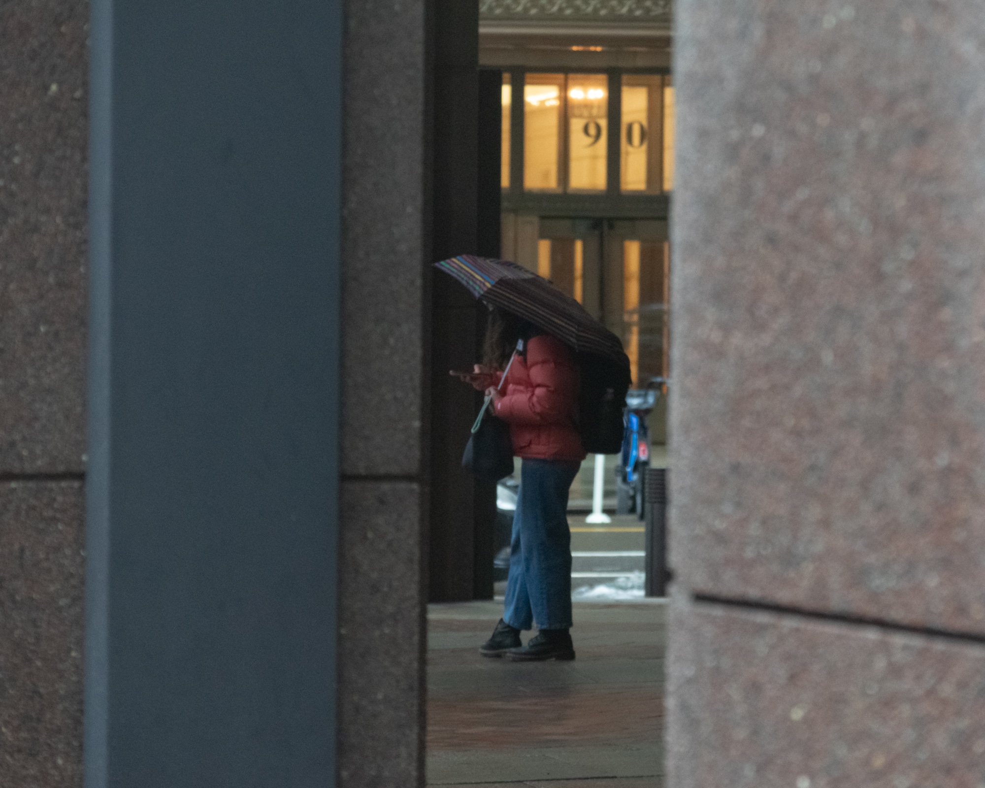 Seen through a narrow gap between two stone building columns, a person in a red puffer jacket and blue jeans stands under a striped umbrella on a wet sidewalk, looking down at a phone. Behind them, a lit lobby entrance shows the building number 90 above glass doors.