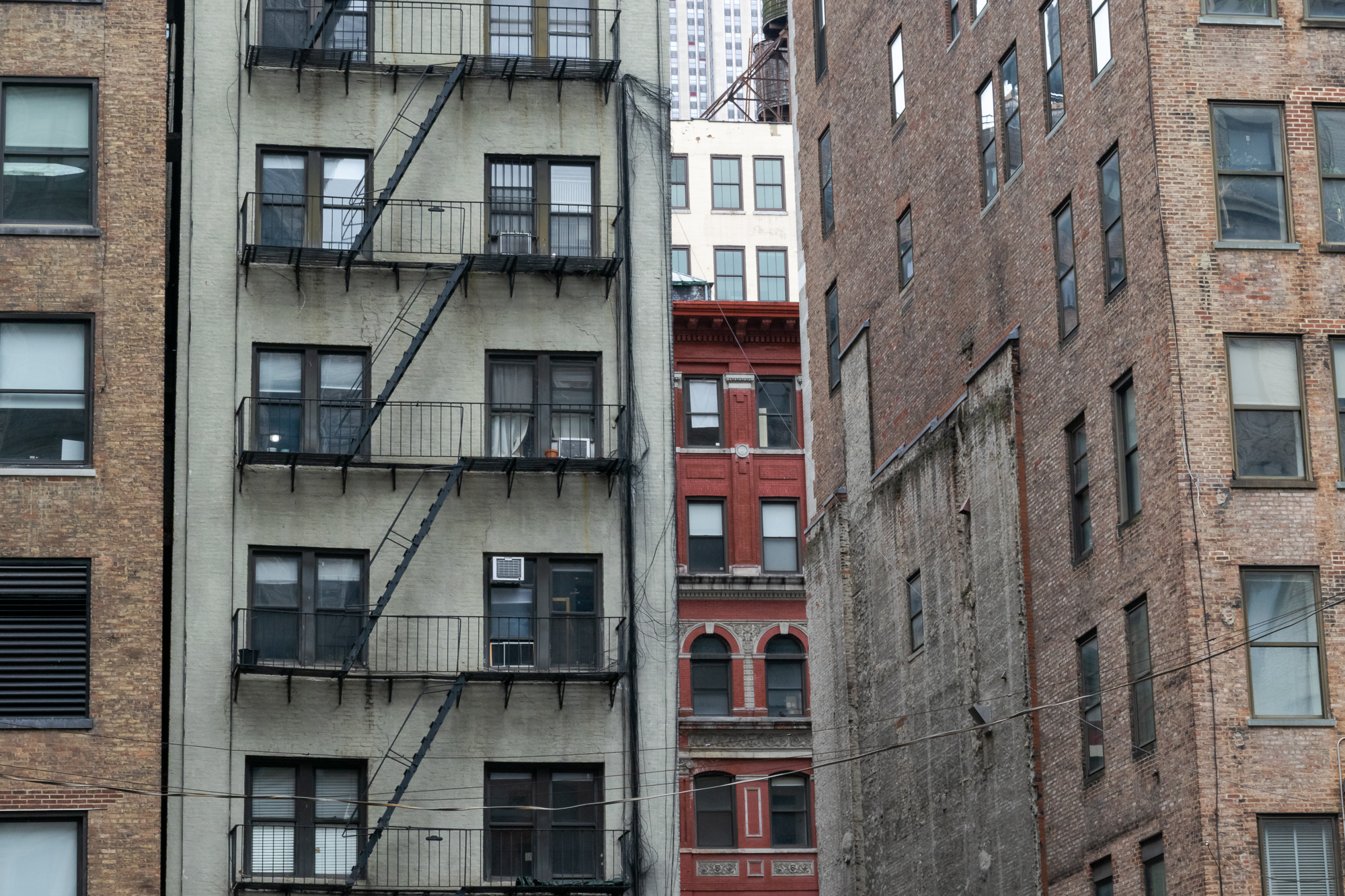 A tight telephoto view of Manhattan building backs: a pale painted brick tenement with a black zigzag fire escape and window air conditioners on the left, a narrow gap revealing a red terracotta facade with arched windows in the center, and a weathered brown brick wall with scarred stucco on the right.