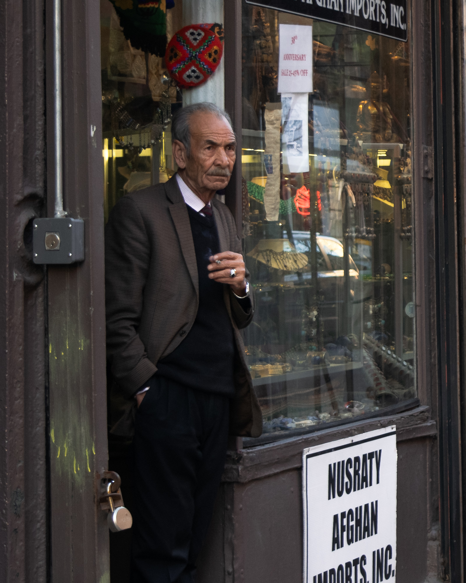 An older man with a gray mustache stands in the doorway of a storefront, holding a lit cigarette in his ringed right hand with his left hand in his trouser pocket. He wears a brown tweed jacket over a black sweater vest and tie, beside a display window crowded with jewelry, brass figures and a sign reading Nusraty Afghan Imports, Inc.