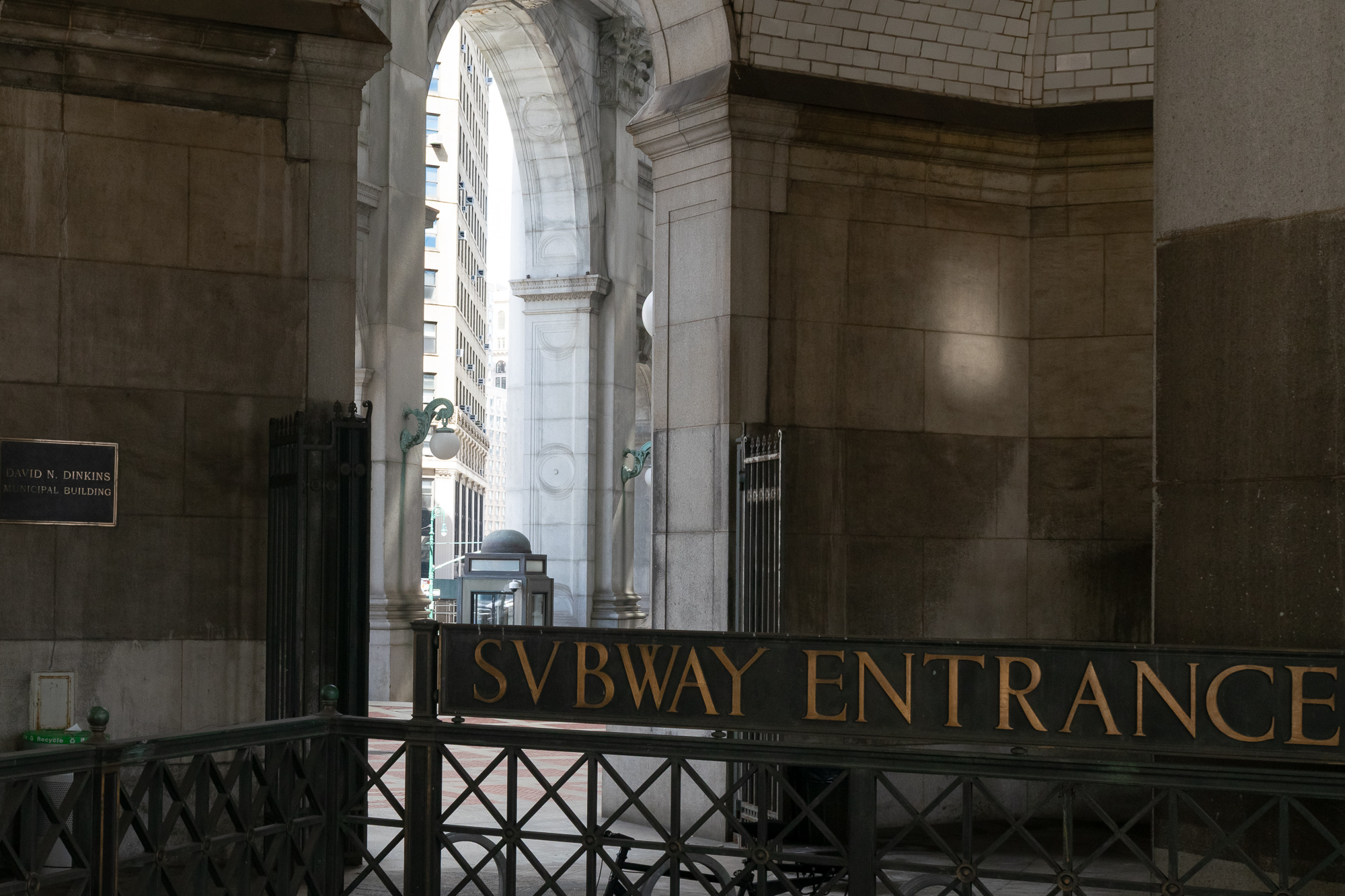 A gilded cast-iron sign reading SVBWAY ENTRANCE, with a V in place of the U, crowns a railing at the top of a subway stair inside the vaulted stone arcade of the David N. Dinkins Municipal Building. Through the archway beyond, daylight falls on a white office tower, a globe lamp on a green bracket, and a small glass guard booth.