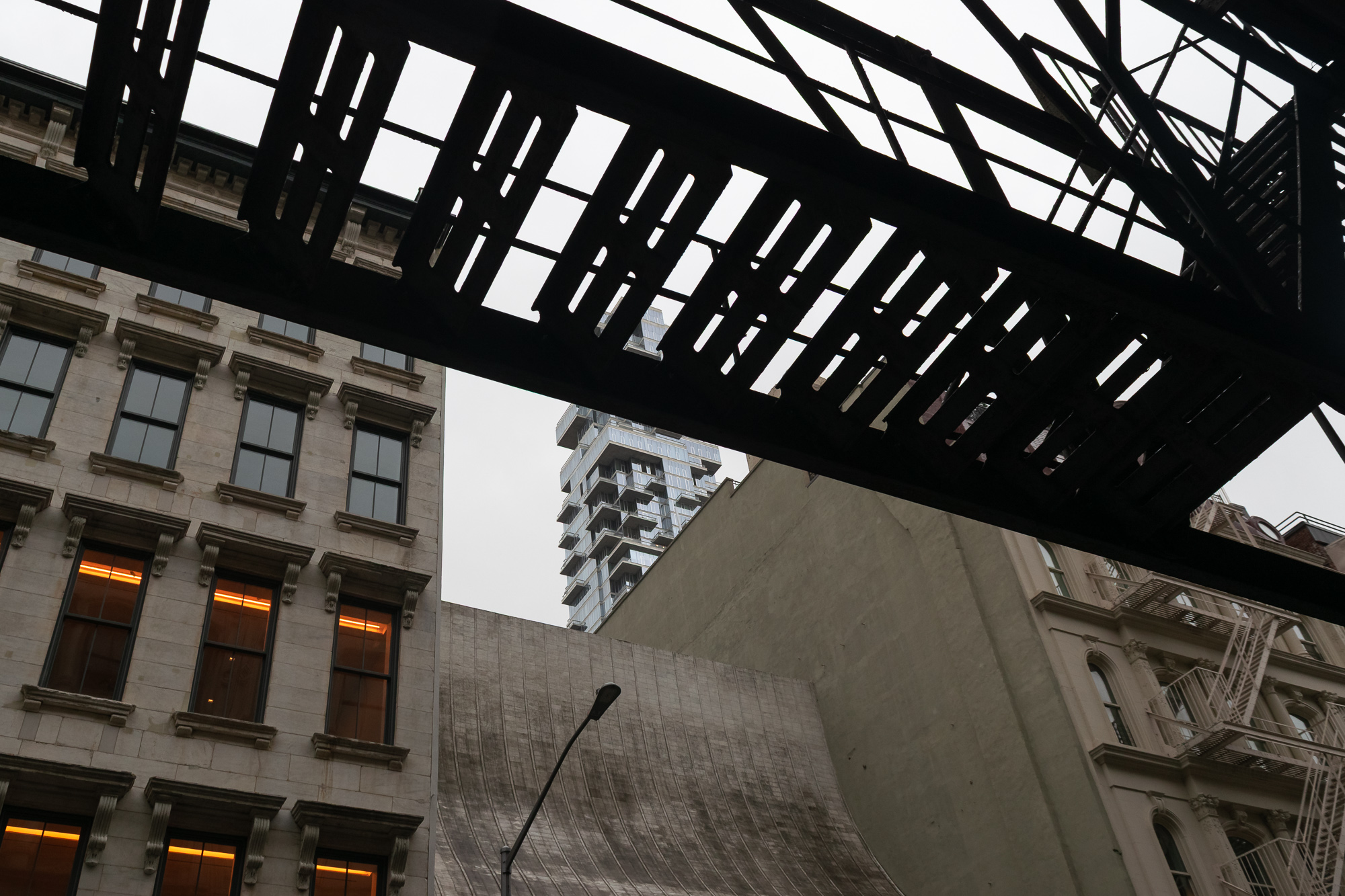 Looking up from a narrow street, a black steel fire escape walkway crosses the top of the frame in silhouette, its slatted treads cut against a flat white sky. Below it sit a pale cast iron building with lit windows on the left, a blank concrete party wall in the center, and a stepped glass residential tower in the distance.