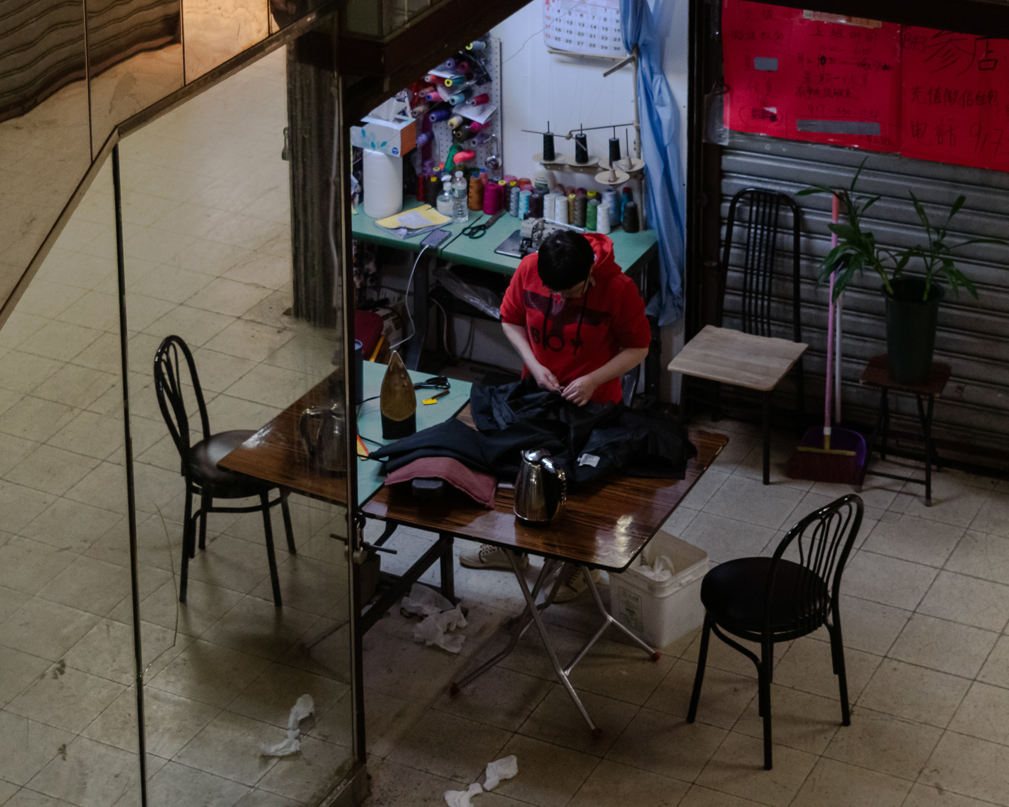 Seen from above, a tailor in a red hooded sweatshirt bends over a black garment at a folding table, surrounded by an iron, a steel kettle and racks of thread spools on a pegboard. A mirrored column at left reflects the tiled floor and empty bentwood chairs.