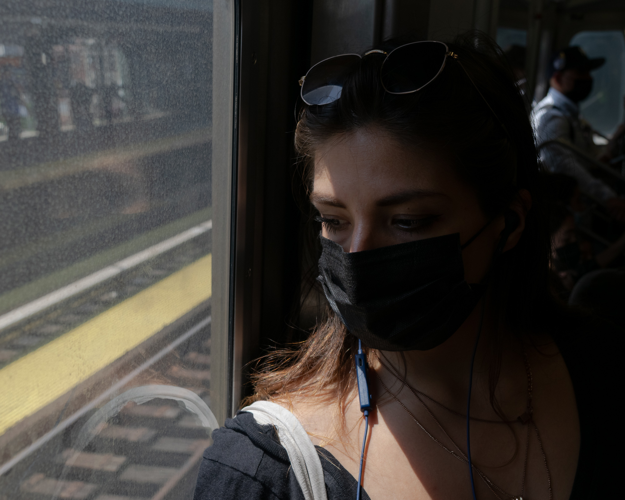 A young woman with dark hair, sunglasses pushed up on her head and a black face mask, leans against a train window as sunlight falls across her eyes and cheek. Through the glass, a station platform with a painted yellow safety stripe is visible, and other masked passengers sit in shadow behind her.