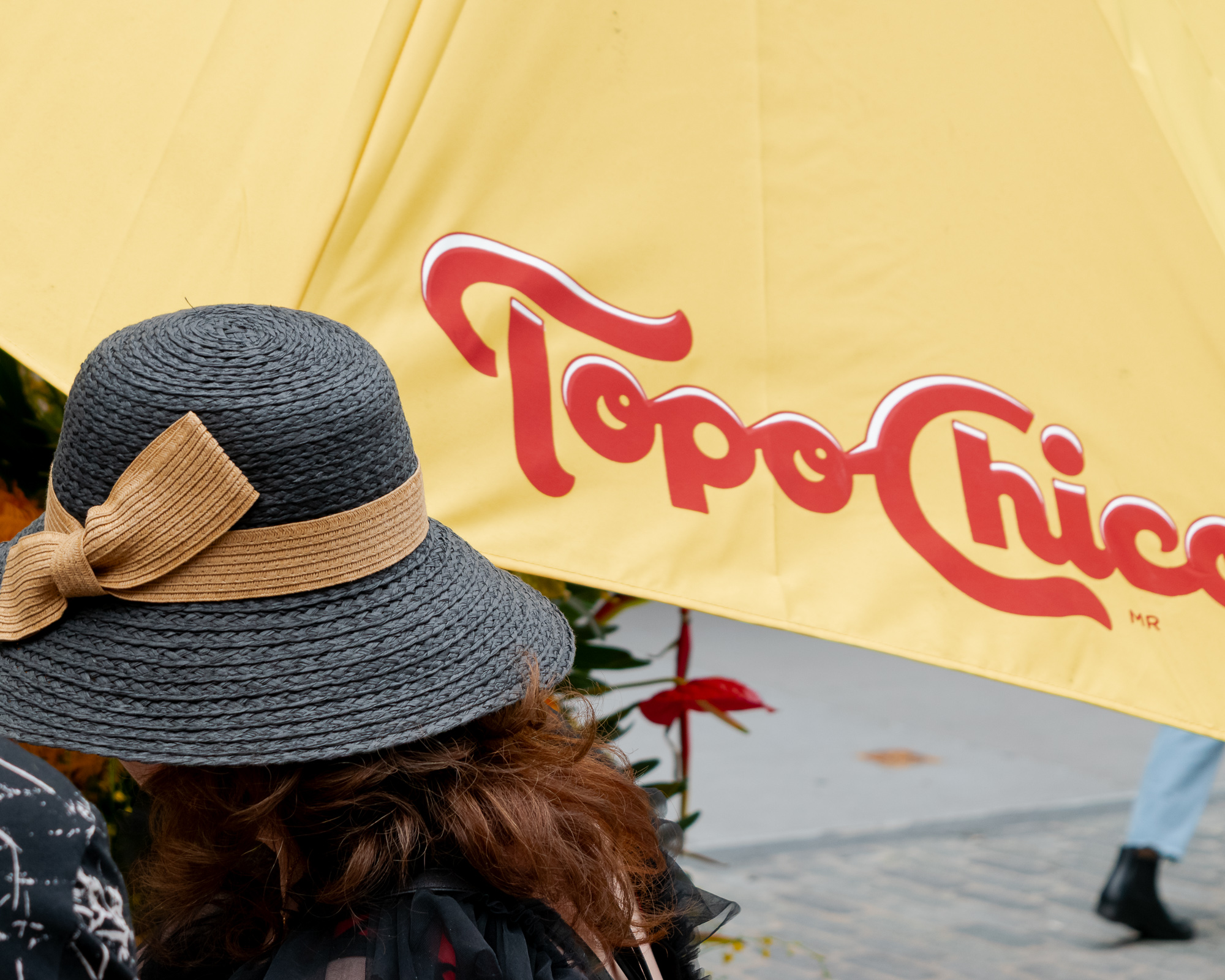 A woman seen from behind wears a wide-brimmed gray straw hat with a tan woven bow, her auburn hair falling below the brim. Behind her, a large yellow patio umbrella fills the frame with the red script Topo Chico logo, and a pedestrian in pale blue jeans and black boots walks across the pavement at right.