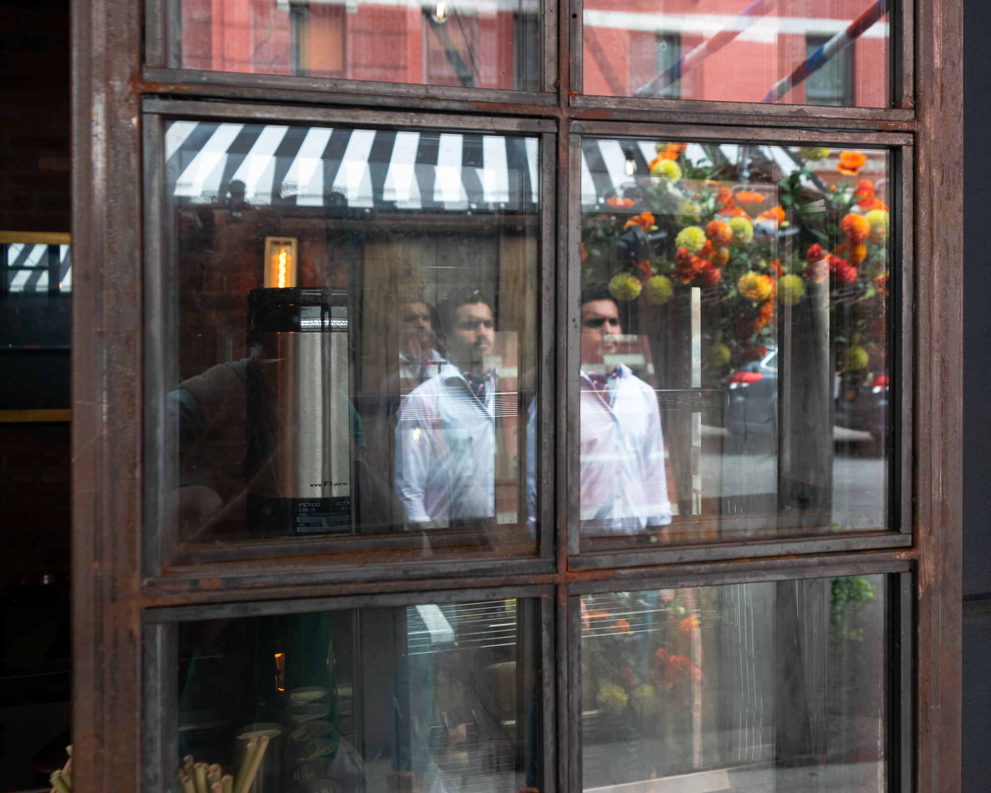 A rusted steel-framed cafe window in which two men in white shirts appear twice, their reflections repeated across adjacent panes alongside a black and white striped awning, a red brick facade, and hanging baskets of orange and yellow flowers. Behind the glass a stainless steel Fetco coffee dispenser and a glowing filament bulb are visible inside the shop.