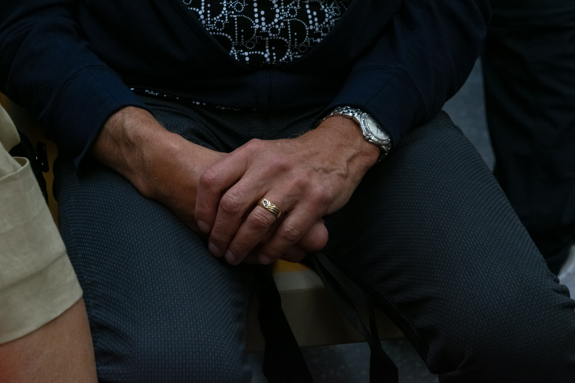 Close-up of a seated man's clasped hands resting on his knees, wearing a gold band on one ring finger and a steel bracelet watch on the left wrist. He wears a navy sweatshirt over a black and white patterned shirt and dark gray textured trousers, with another passenger's tan sleeve at the left edge.