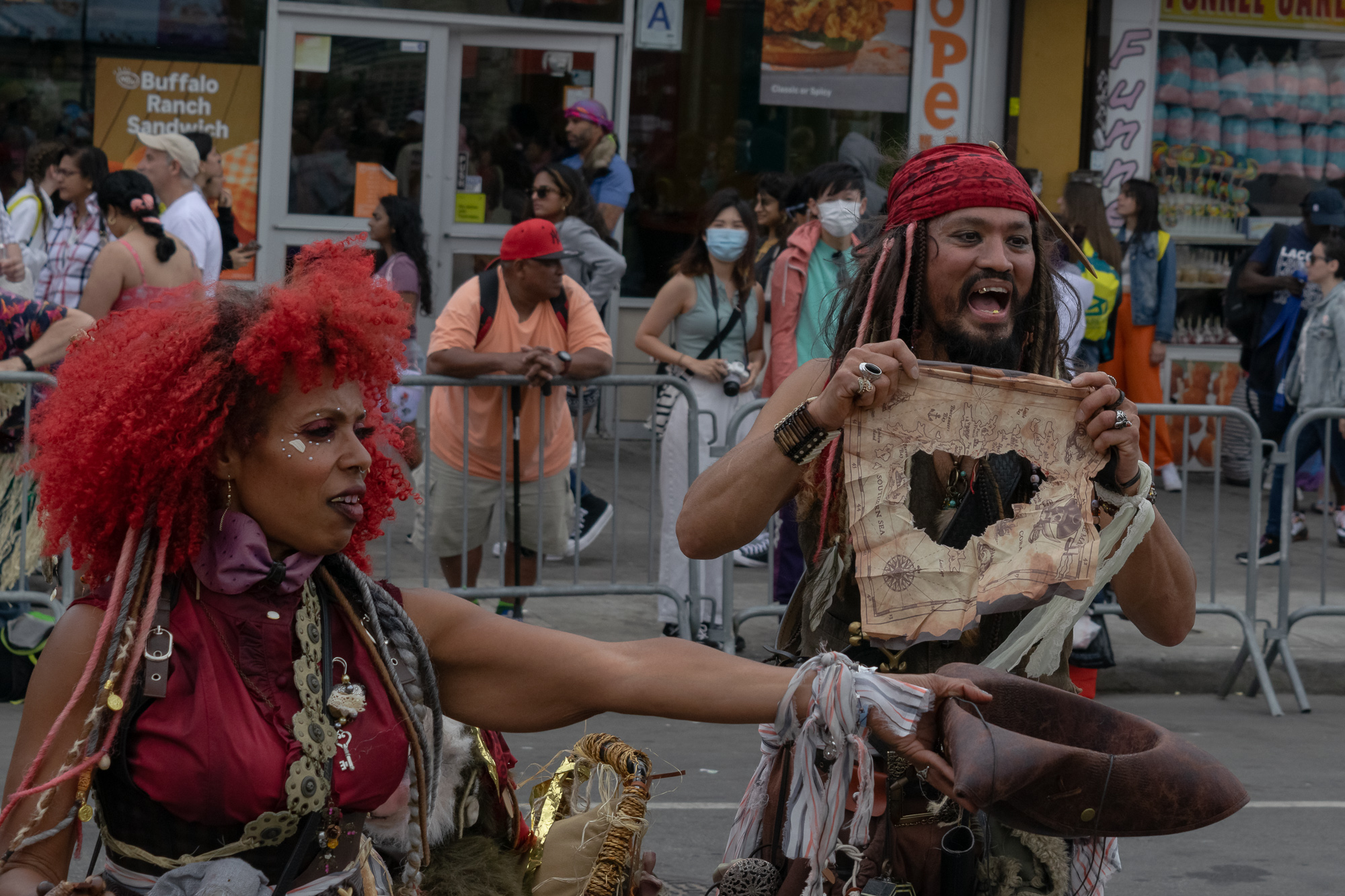 Two costumed parade performers on a Brooklyn street: a woman with a large red curly wig, white face paint under one eye and a purple bow tie reaching out with one arm, and a man in a red bandana with beaded dreadlocks holding up a torn parchment treasure map and a leather tricorn hat. Behind them, spectators stand along metal barricades in front of a fast food window and a funnel cake and cotton candy stand.