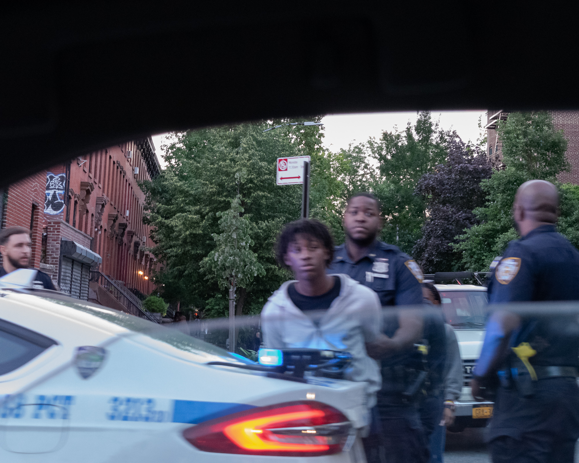 Viewed through a car windshield with the dark roofline framing the top of the frame, a young man in a white hooded sweatshirt is held by a uniformed NYPD officer on a tree-lined street, while another officer stands blurred in the foreground at right and a marked police car with lit taillight and roof lights fills the lower left. A red brick rowhouse with graffiti and a no-parking sign line the background.