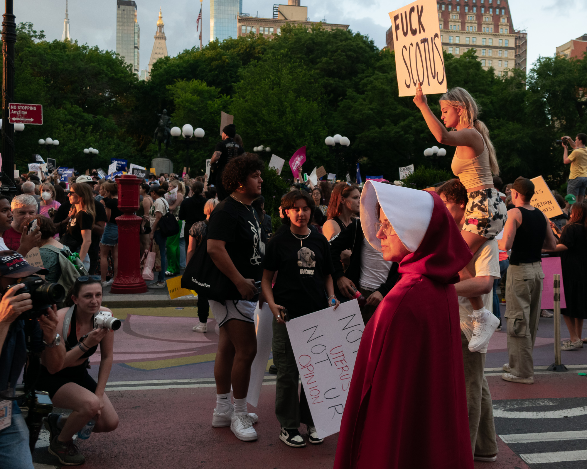 A dense crowd fills a street at Union Square in New York City during an abortion rights protest, with a woman in a red cloak and white bonnet in the right foreground and a blonde woman seated on someone's shoulders raising a hand-lettered sign reading FUCK SCOTUS above the trees.