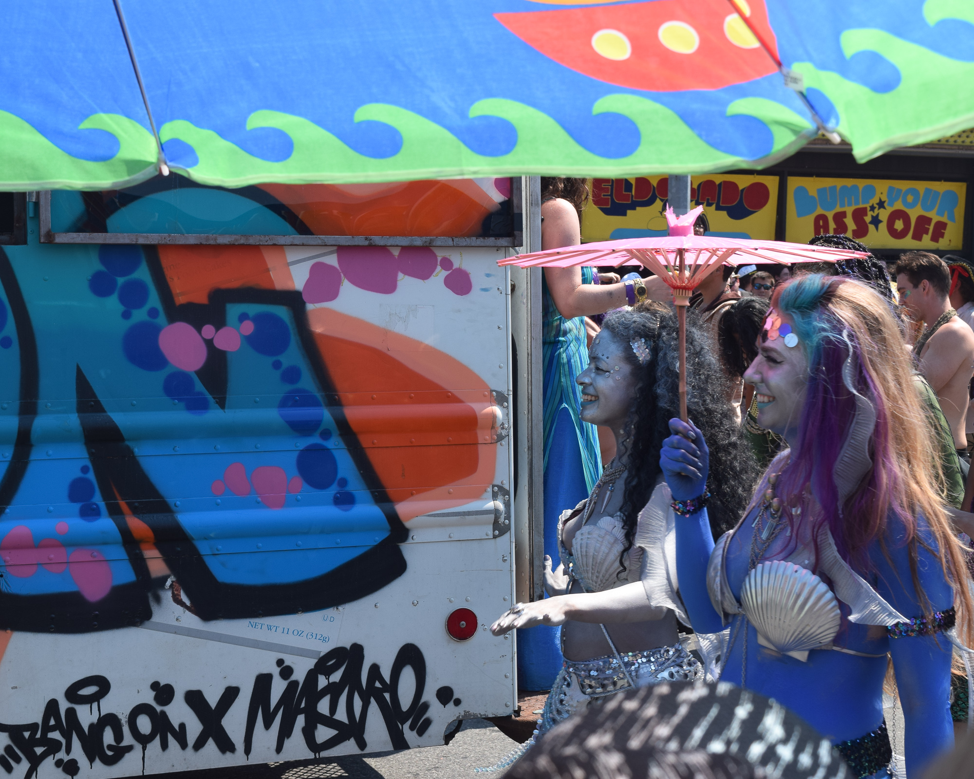 Two women in blue body paint, shell bras and glitter walk past a graffiti-covered box truck under a wave-patterned awning at the Coney Island Mermaid Parade, one holding a pink paper parasol, with a yellow Eldorado bumper car sign reading 'Bump Your Ass Off' behind them.