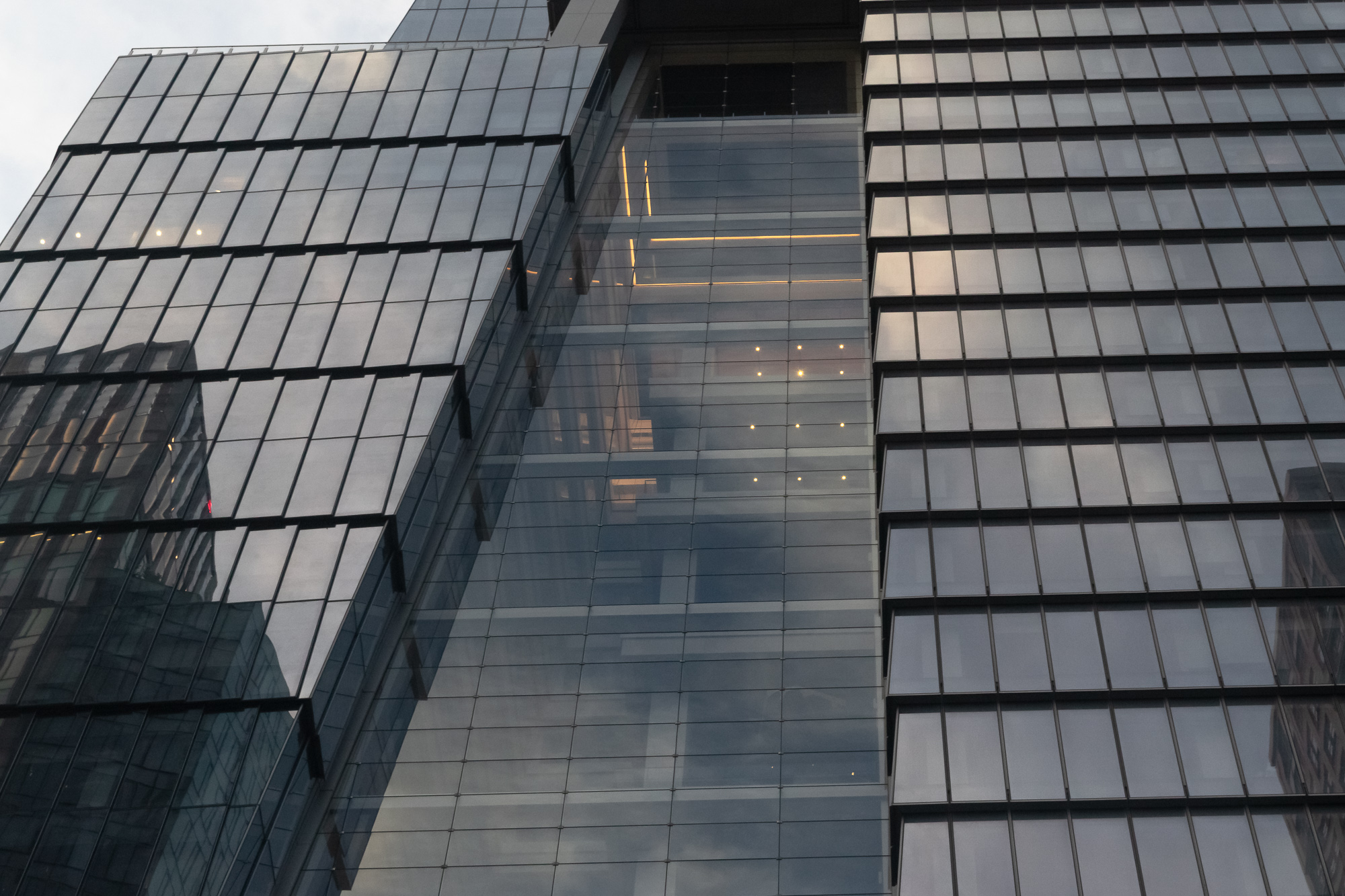 Looking up at a modern skyscraper's glass curtain wall, where two stepped, angled facades flank a narrow recessed slot of darker glazing. Pale evening sky and neighboring buildings reflect across the panels, and a scatter of small warm ceiling lights and a horizontal amber strip glow from inside the upper floors.