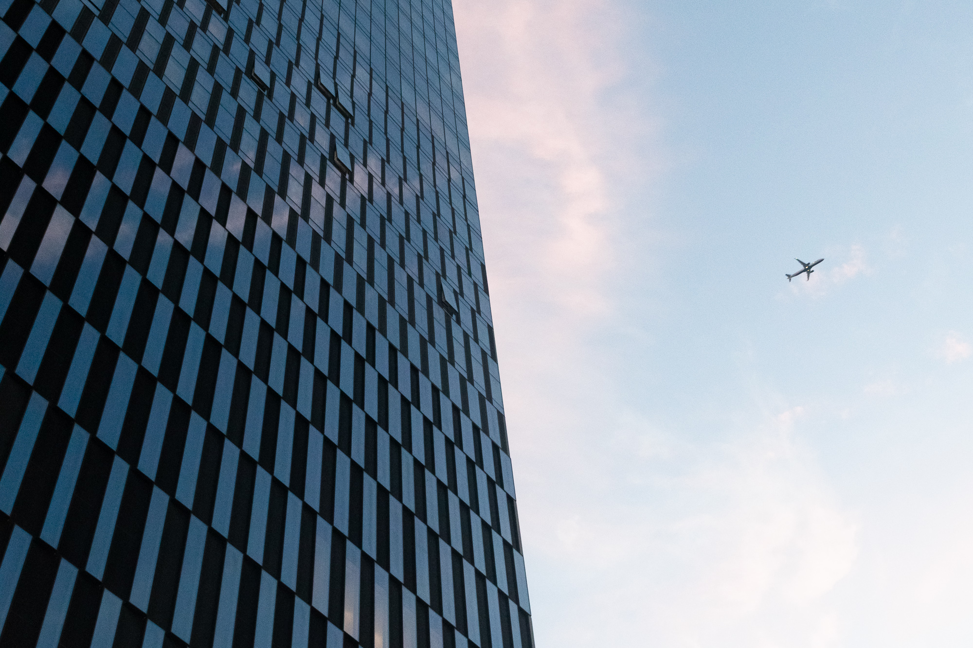 Looking up at the corner of a skyscraper clad in a staggered checkerboard of dark panels and blue reflective glass, its edge cutting diagonally across a pale sky. A commercial jet, seen from below with wings spread, flies in the open sky to the right.