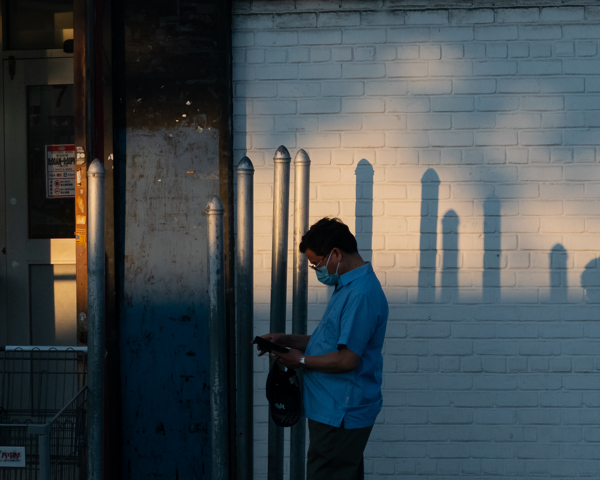 A man in a light blue short-sleeved shirt and surgical mask stands in profile outside a storefront, looking down at a phone while holding a black cap in his other hand. Behind him, late sun throws long shadows of steel sidewalk bollards across a painted white brick wall.