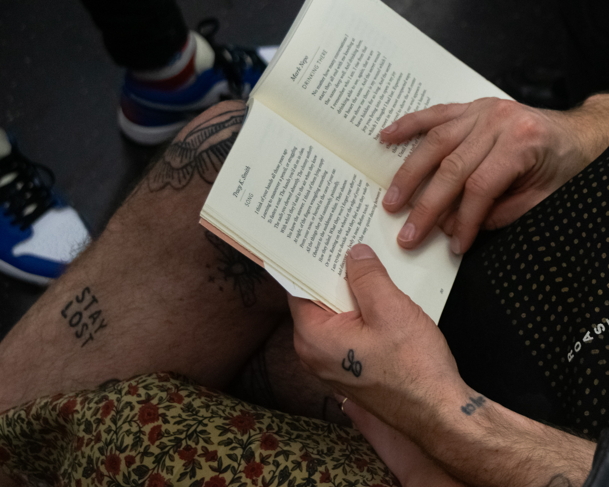 Overhead view of a seated person on a subway holding open a poetry anthology, with pages showing poems by Mark Nepo and Tracy K. Smith; their bare thigh carries a 'STAY LOST' tattoo and their hand bears a small looped symbol. Across the aisle, a pair of red, white and blue sneakers rests on the train floor.