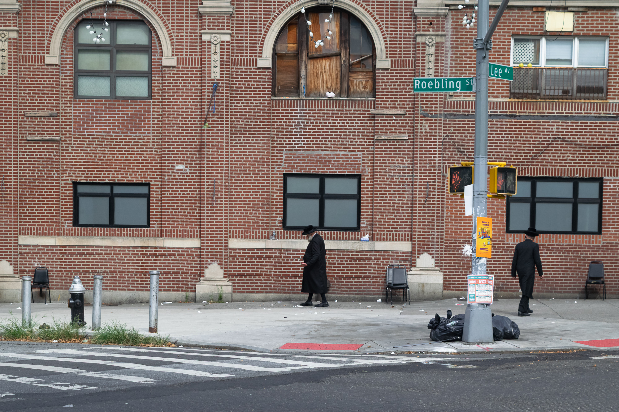 Two men in long black coats and wide-brimmed hats walk in opposite directions along the sidewalk in front of a red brick building with boarded-up arched windows, at the corner of Roebling Street and Lee Avenue. A pedestrian signal shows a red hand, and black trash bags sit at the base of the traffic pole.