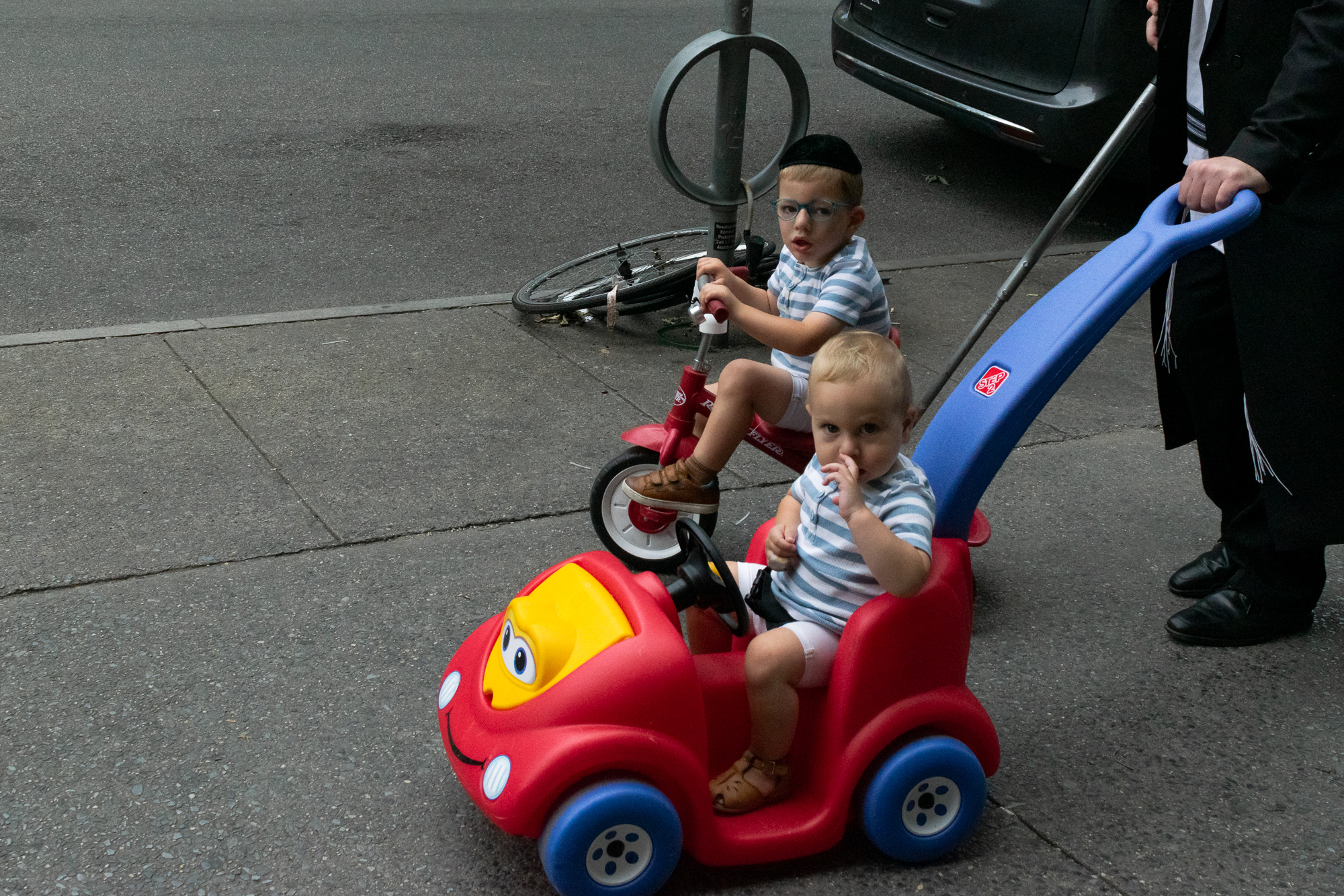 A blond toddler in a blue-and-white striped shirt sits in a red plastic push car with a yellow face and blue wheels, fingers at his mouth, while an older boy in glasses and a black yarmulke sits on a red Radio Flyer tricycle just behind him. A man in a black suit with visible tzitzit grips the car's blue push handle at the right edge of the frame.