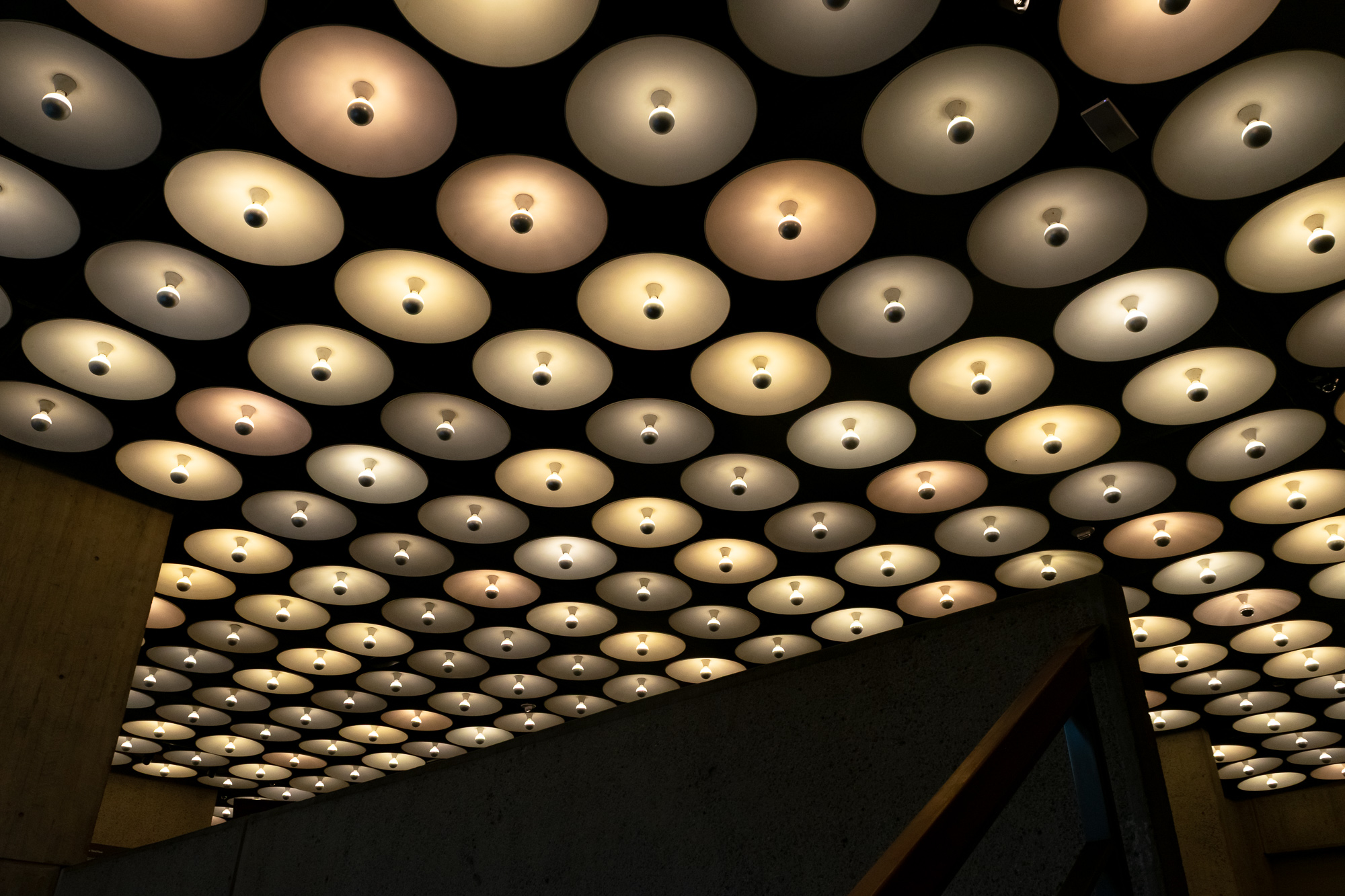 Looking up at a dark ceiling filled with rows of wide, shallow dome light fixtures, each with a bare bulb at its center, glowing in warm white, amber and faintly pink tones. The grid recedes toward the upper right and is cut off at the lower right by the dark diagonal edge of a concrete wall and a wood-topped railing.