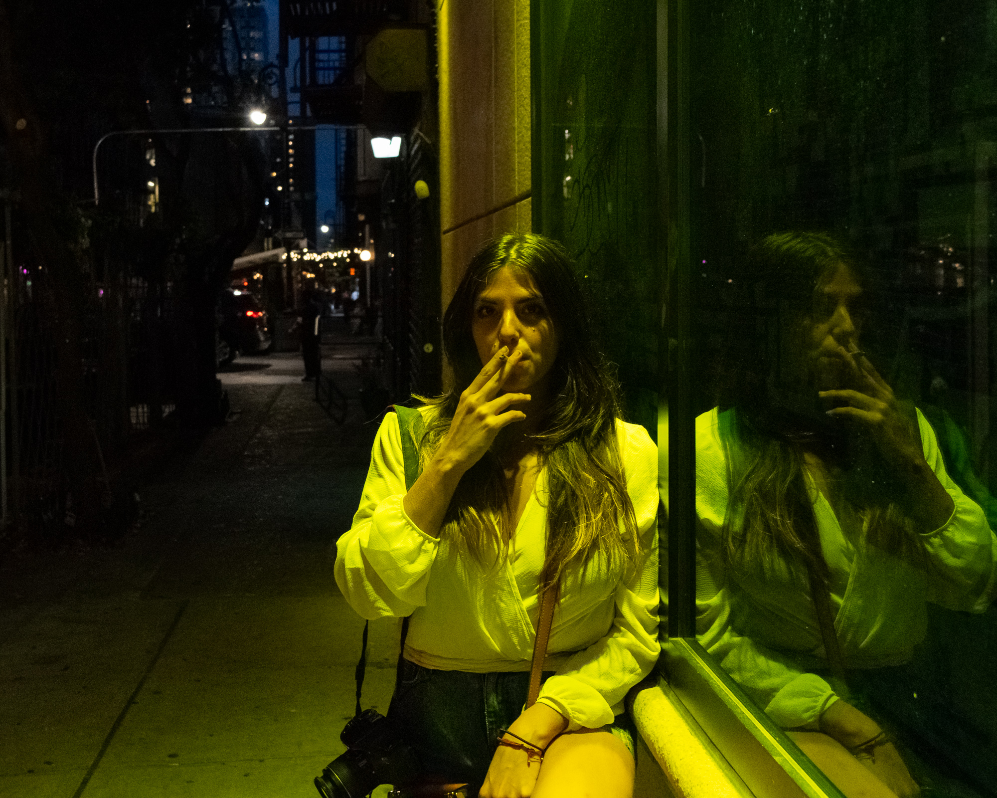 A woman in a white blouse sits against a tinted green storefront window at night, raising a cigarette to her lips, her face lit yellow by overhead light. Her reflection repeats in the glass to the right, while behind her a dark sidewalk recedes toward fire escapes and distant string lights.