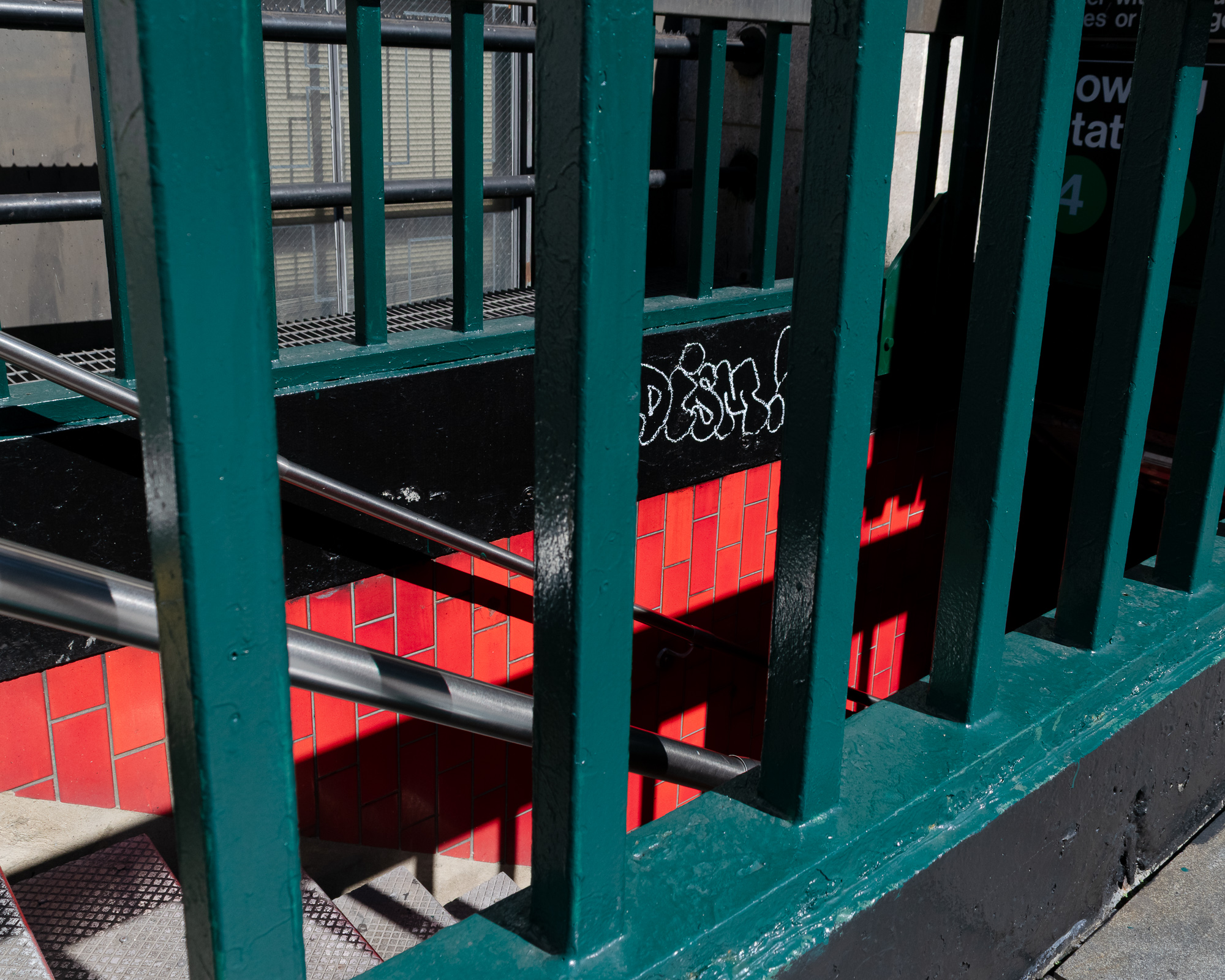 Looking down through thick green-painted vertical railings into a New York City subway entrance, where steel handrails descend past bright red brick-pattern tile walls. A white chalk-outline graffiti tag sits on the black painted wall above the tile, with a station sign partly visible at right.