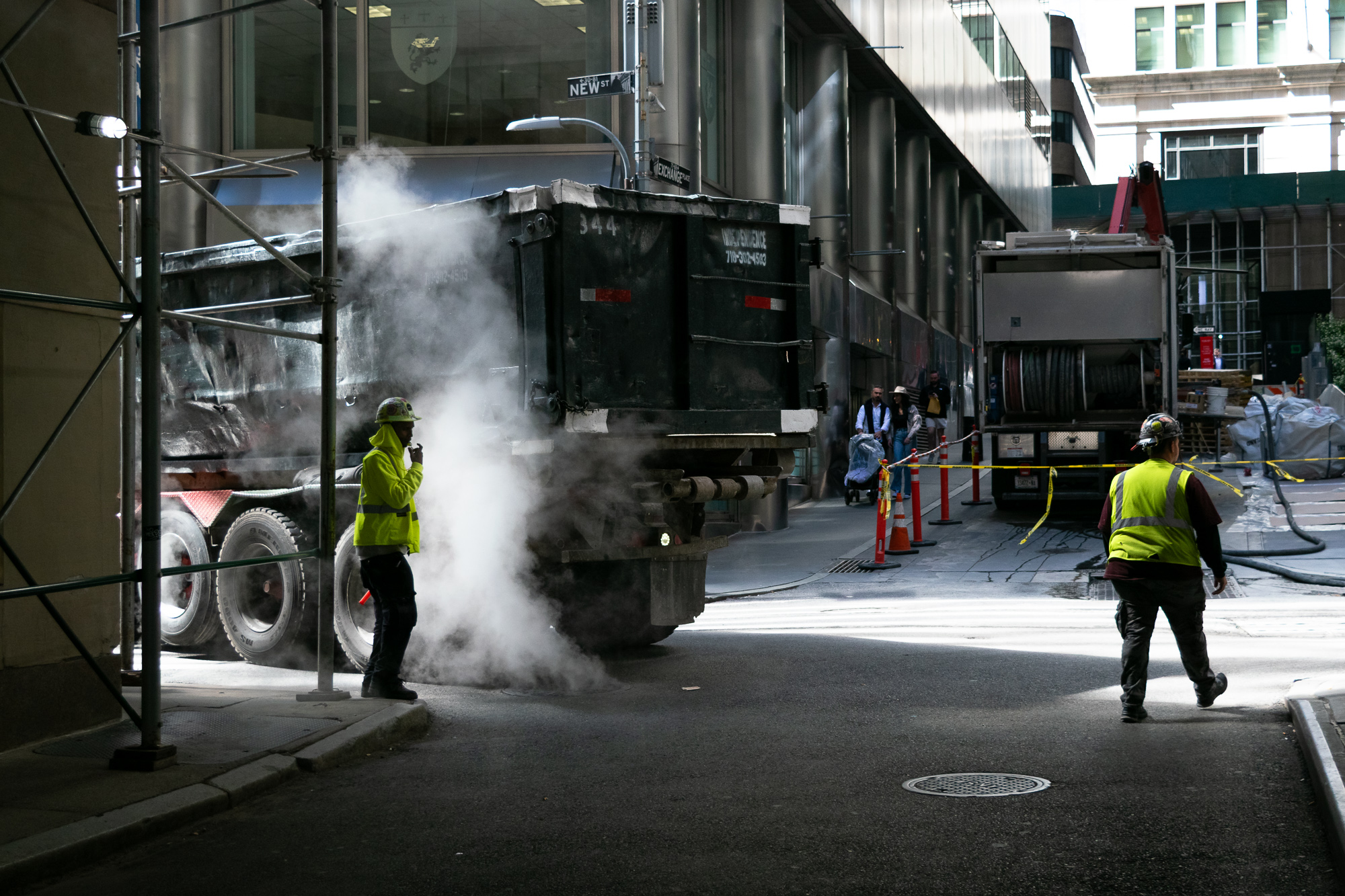 A thick plume of white steam rises around the rear of a black dump truck parked on a narrow Manhattan street, while a worker in a yellow high-visibility jacket and hard hat stands beside it holding a radio. A second worker in a yellow vest and ear-protection helmet walks across the asphalt toward a utility truck strung with yellow caution tape.