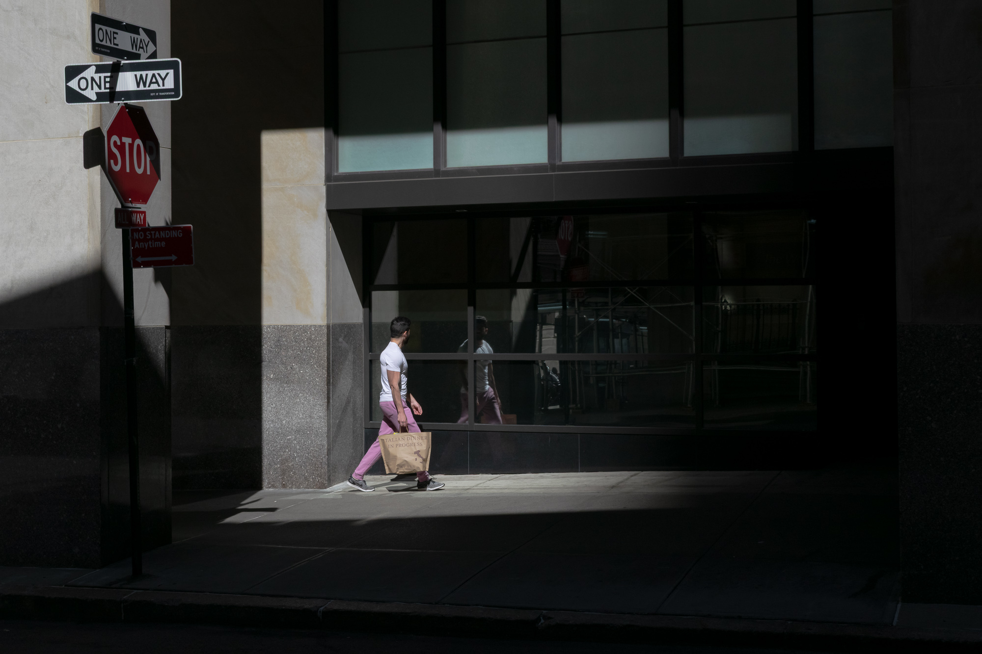 A man in a white t-shirt and pink sweatpants walks along a sunlit strip of sidewalk carrying a paper shopping bag printed with the words Italian Dinner in Progress, his figure mirrored in the dark plate glass of the building beside him. At the left edge, two One Way signs, a stop sign, and a No Standing Anytime sign stack on a single pole in deep shade.