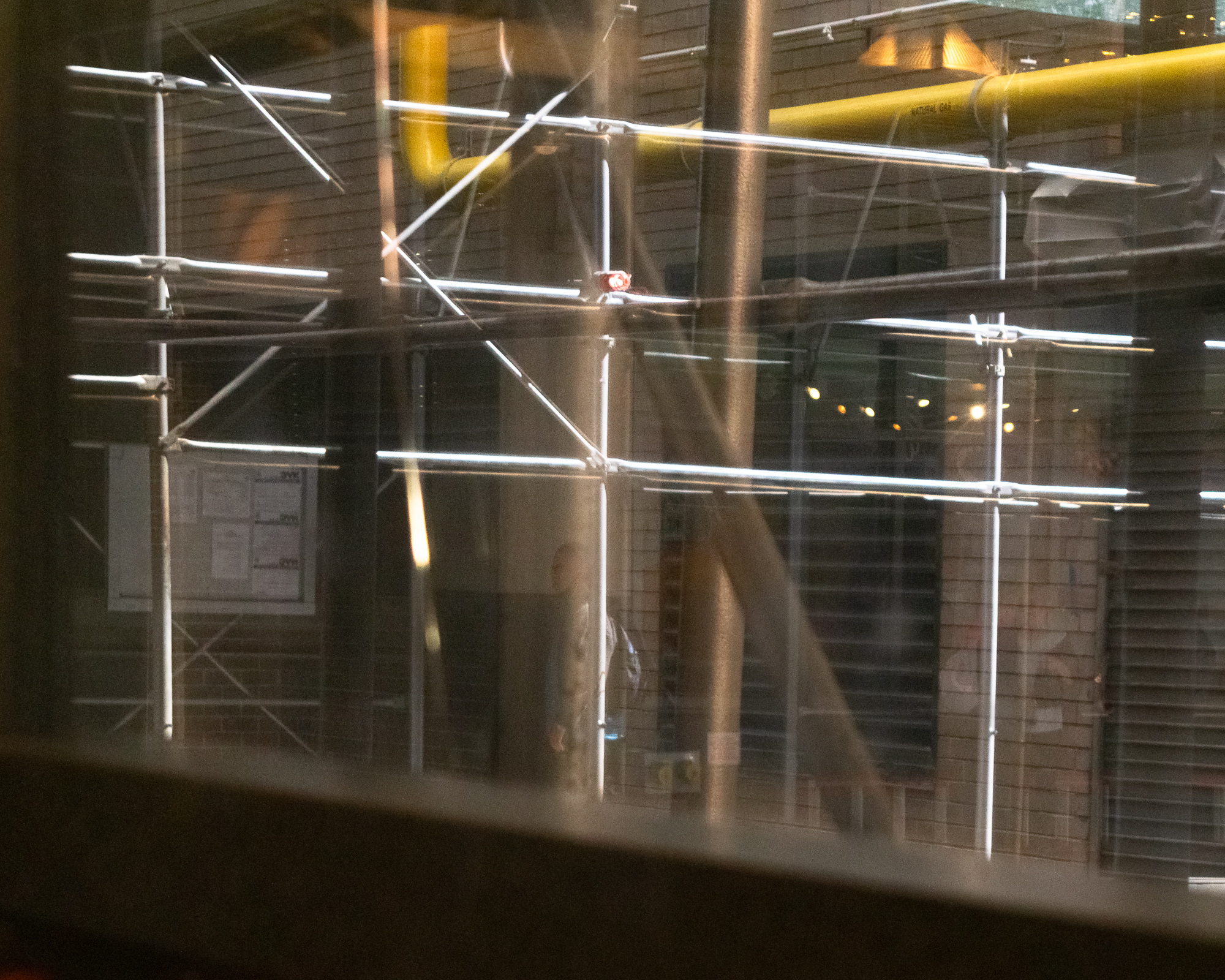 Shot through a window with visible glare and reflections, a lattice of metal scaffolding poles crosses a brick building facade, with a yellow natural gas pipe running horizontally along the upper right and a red light fixture clamped mid-frame.