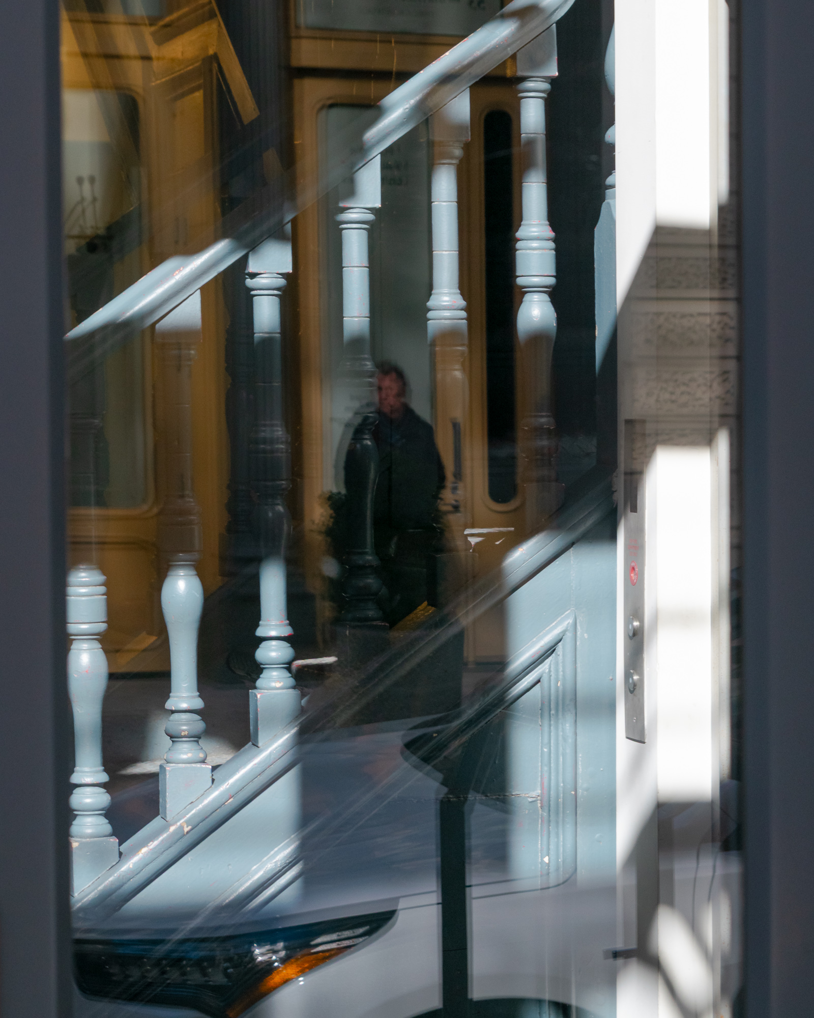 A plate glass window reflects a painted staircase with pale blue turned balusters and a diagonal handrail, layered over the interior of a lobby. Behind the glass, a man in a dark coat stands in a doorway near a tan paneled wall, with a brushed metal call panel and a sunlit white wall at right.