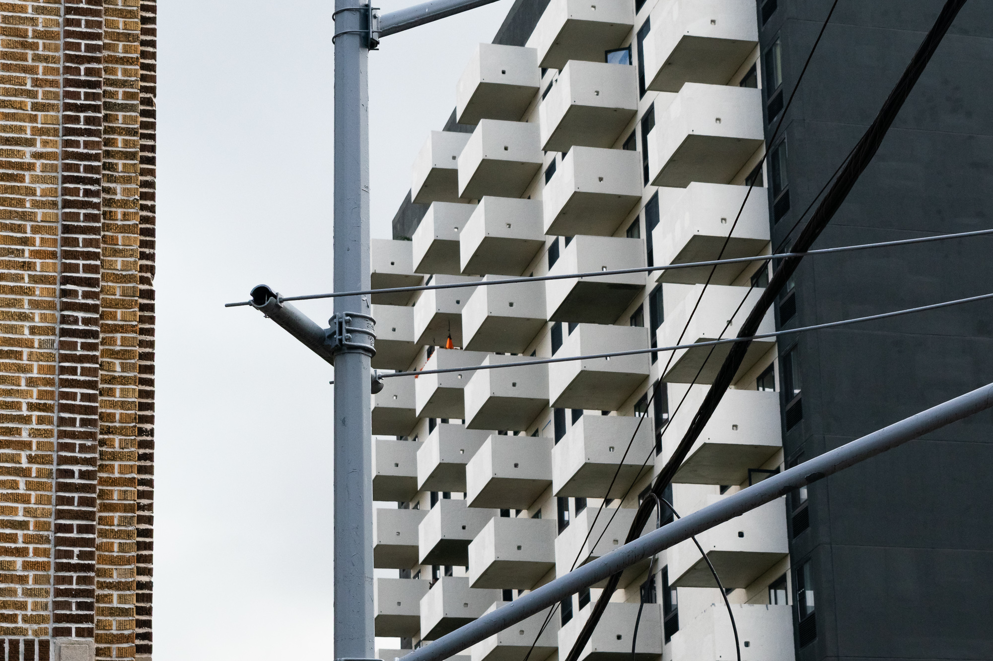 A high-rise apartment tower with rows of cantilevered white concrete balconies rises behind a gray steel utility pole and crossing power lines, with the patterned brick wall of an older building at left against a pale overcast sky.