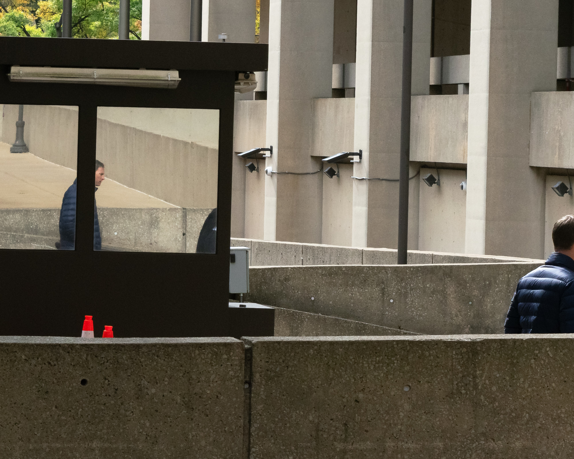 A security booth with dark metal framing and large glass panes reflects a man in a navy puffer jacket walking past a concrete ramp, while at right another man in a navy puffer jacket stands behind a low concrete barrier. Beyond them rise raw concrete columns fitted with floodlights, solar panels and a dome camera.