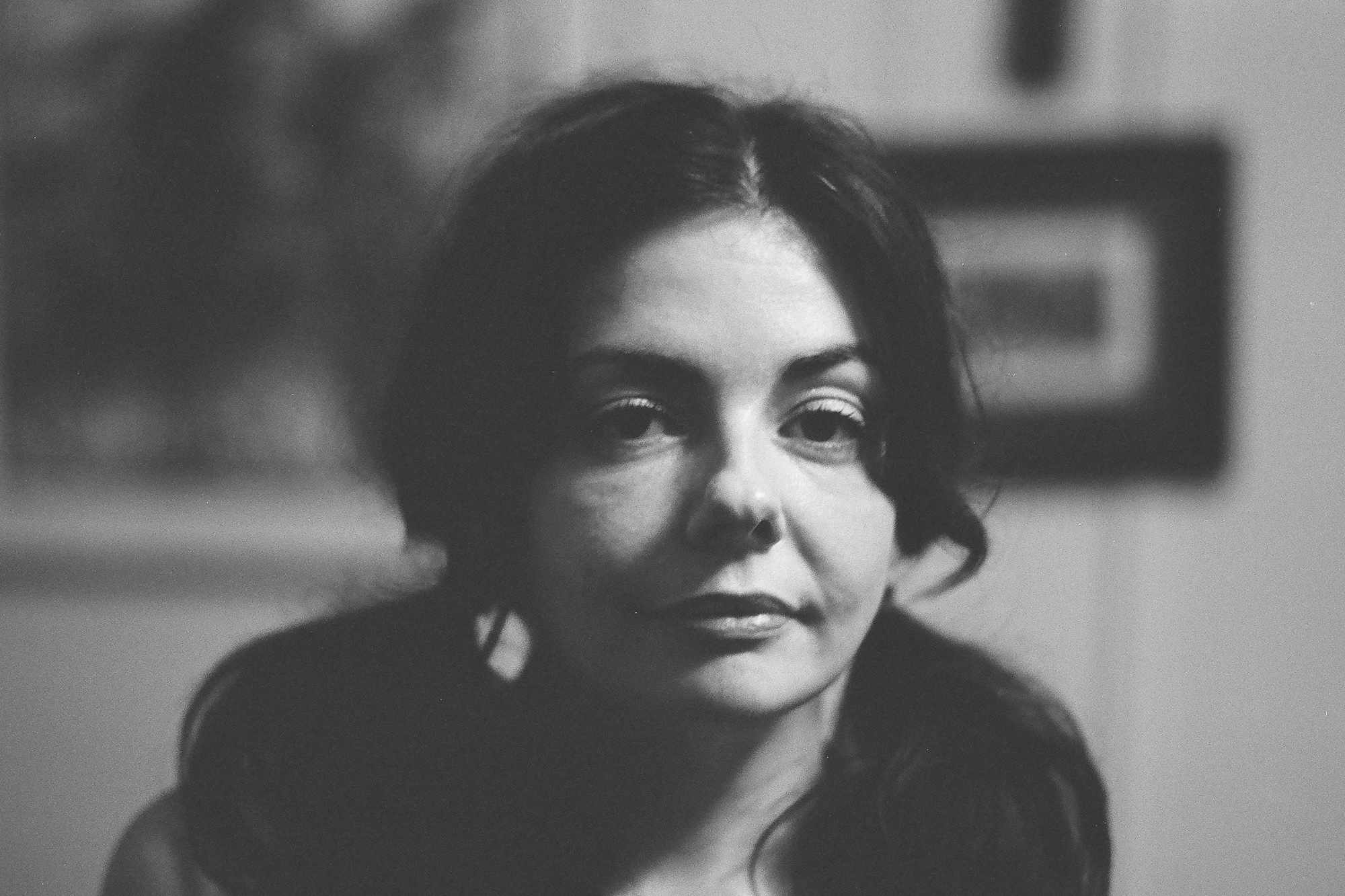 Tight black and white portrait of a dark-haired woman looking just past the camera, her face lit softly from the left, with a blurred framed picture and pale wall behind her.