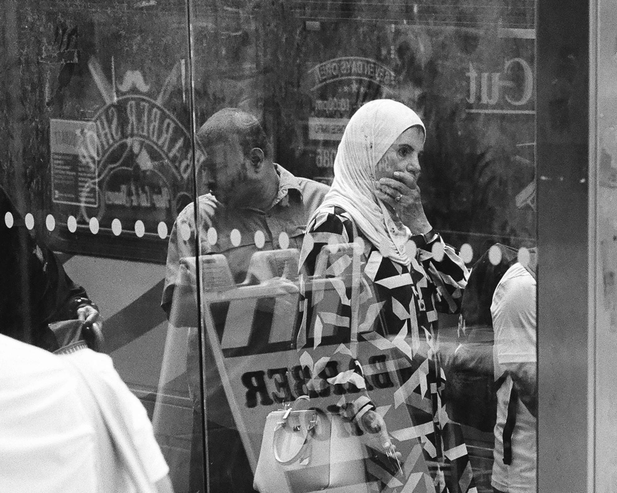 Black and white photograph of a woman in a light headscarf and geometric-print blouse standing behind a plate glass storefront, one hand raised to her mouth, holding a pale handbag. Reversed lettering reading BARBER and CUT floats across the glass along with the reflection of a man in a short-sleeved shirt looking down.