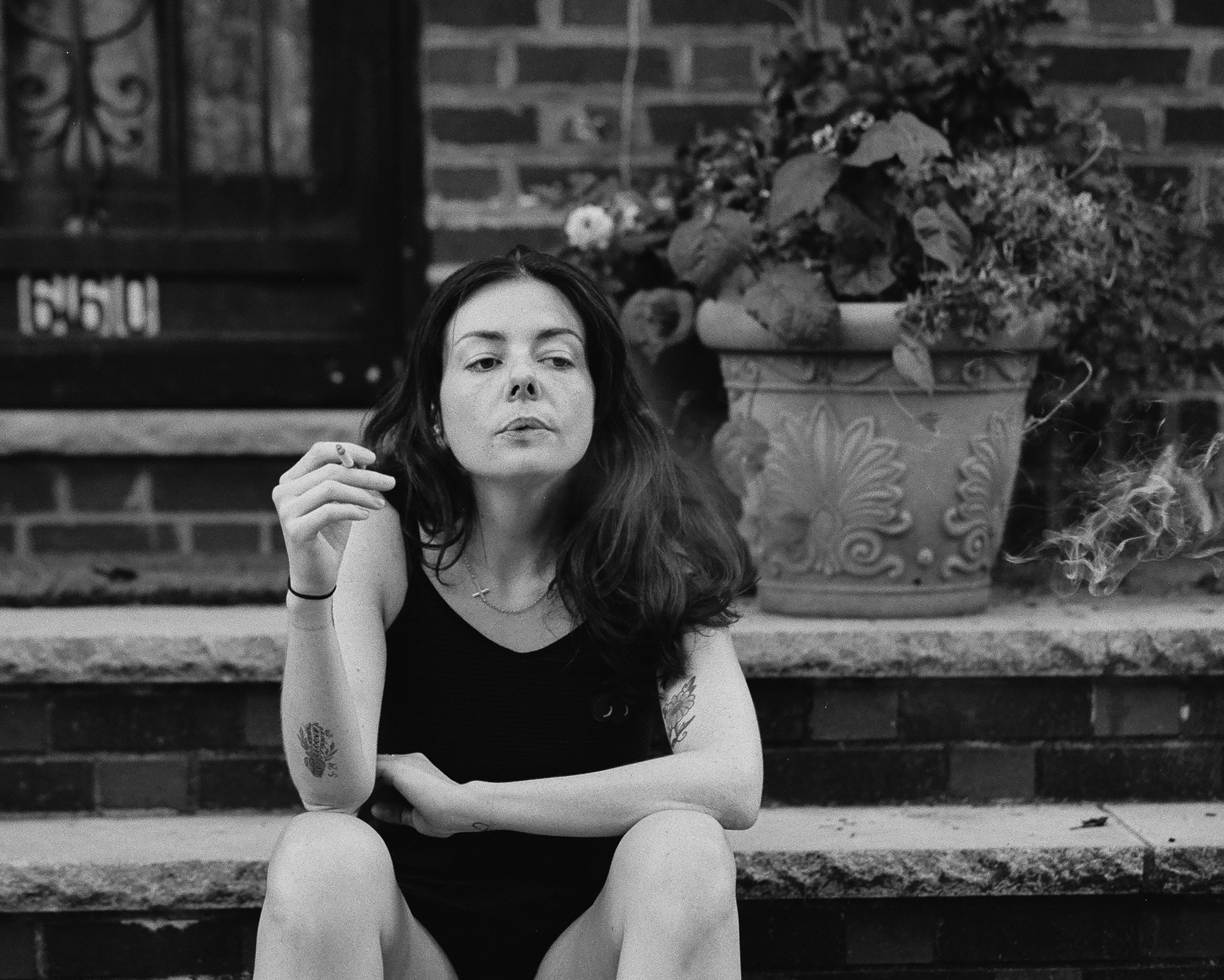 Black and white photograph of a young woman with long dark hair sitting on stone steps outside a brick building, holding a lit cigarette in her raised right hand and exhaling. She wears a dark sleeveless top and a thin cross necklace, with tattoos on her forearm and upper arm, and a large decorative planter of trailing flowers sits on the step behind her.