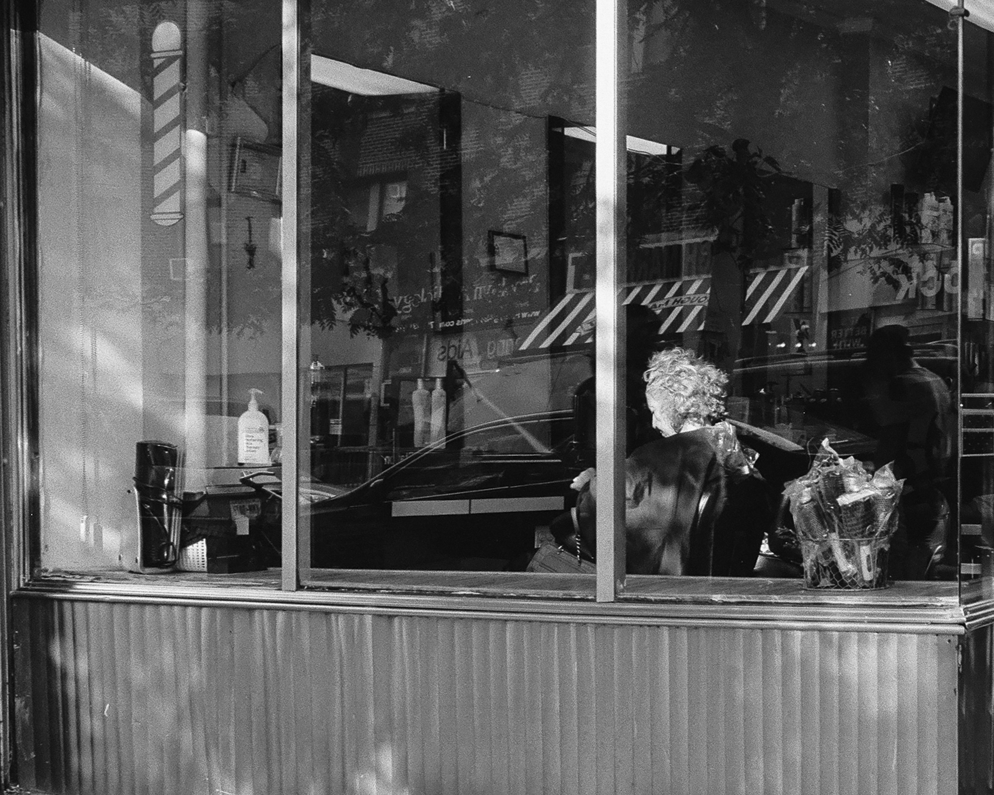 Black and white photograph looking through a large barbershop window where a client with curly white hair sits in a chair wearing a dark cape, while reflections of parked cars, street trees and a brick building overlay the glass. A barber pole decal, a pump bottle of conditioner and a wire basket of wrapped goods sit along the window ledge above corrugated metal siding.