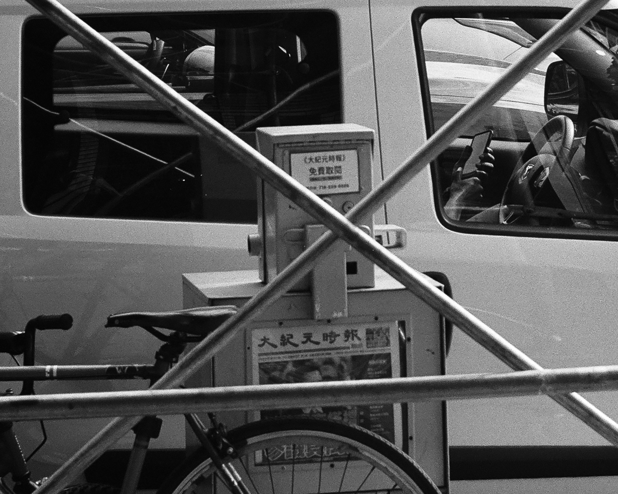 Black and white street photograph looking through crossed metal scaffolding poles at a parked van and car; a driver inside holds up a smartphone, and a newspaper vending box with Chinese characters stands between the vehicles beside a parked bicycle saddle and wheel.